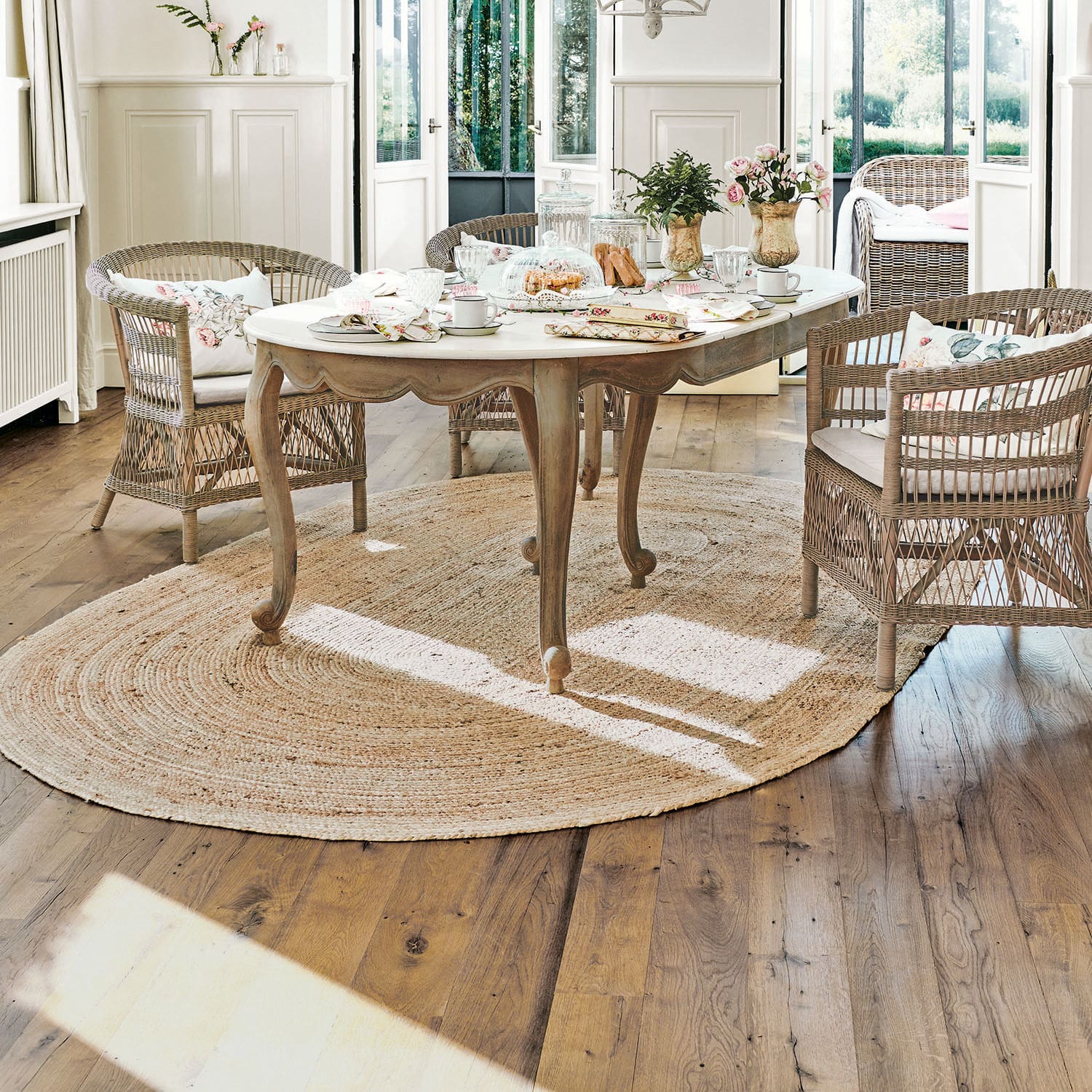 Vloerkleed Ausone Lichte houten tafel met wit blad op ronde jute vloerkleed in lichte kamer met rotan stoelen