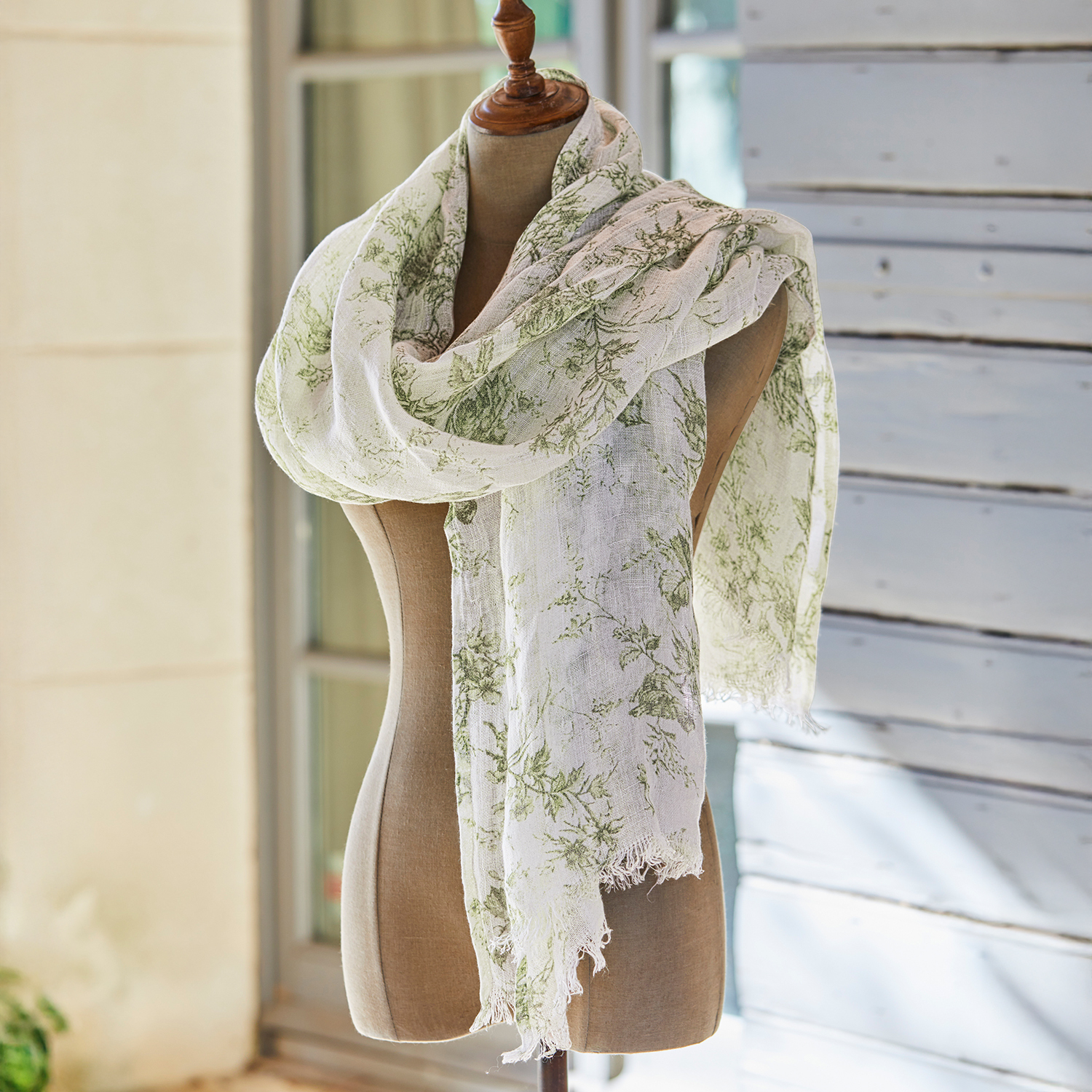 Écharpe légère à motif floral vert sur fond crème, présentée dans un jardin avec panier en osier