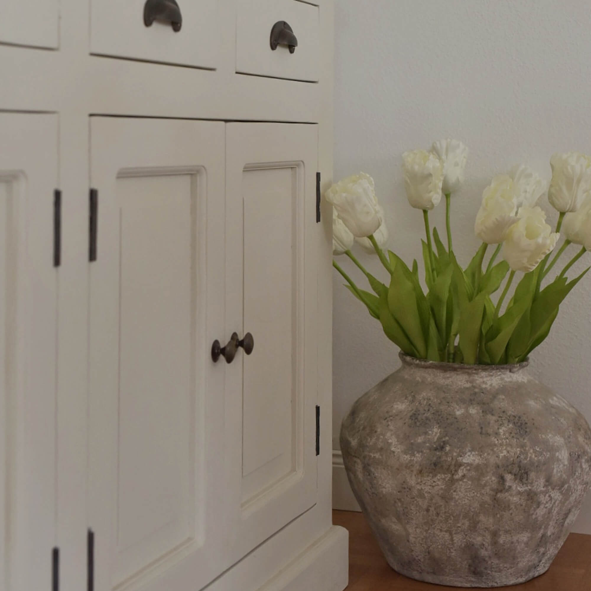 Grote grijze keramische vaas met witte tulpen naast een witte houten kast