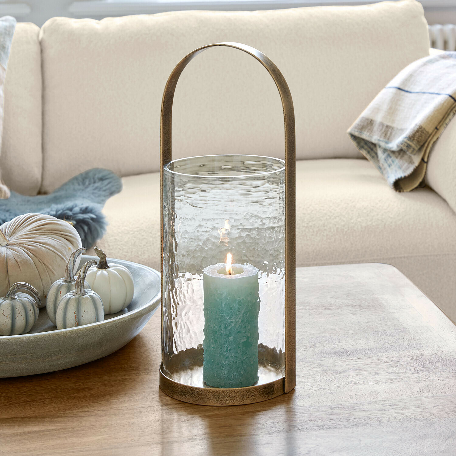 Windlicht Omirah Glazen lantaarn met messing handvat en blauwe kaars op houten tafel in woonkamer