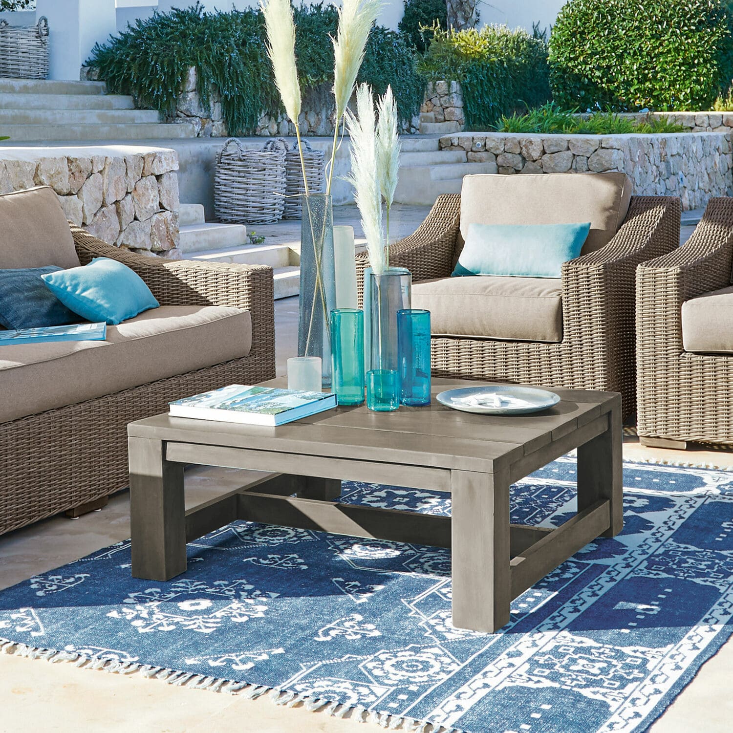 Table basse en bois gris sur tapis bleu à motifs, entourée de fauteuils en osier avec coussins bleus.