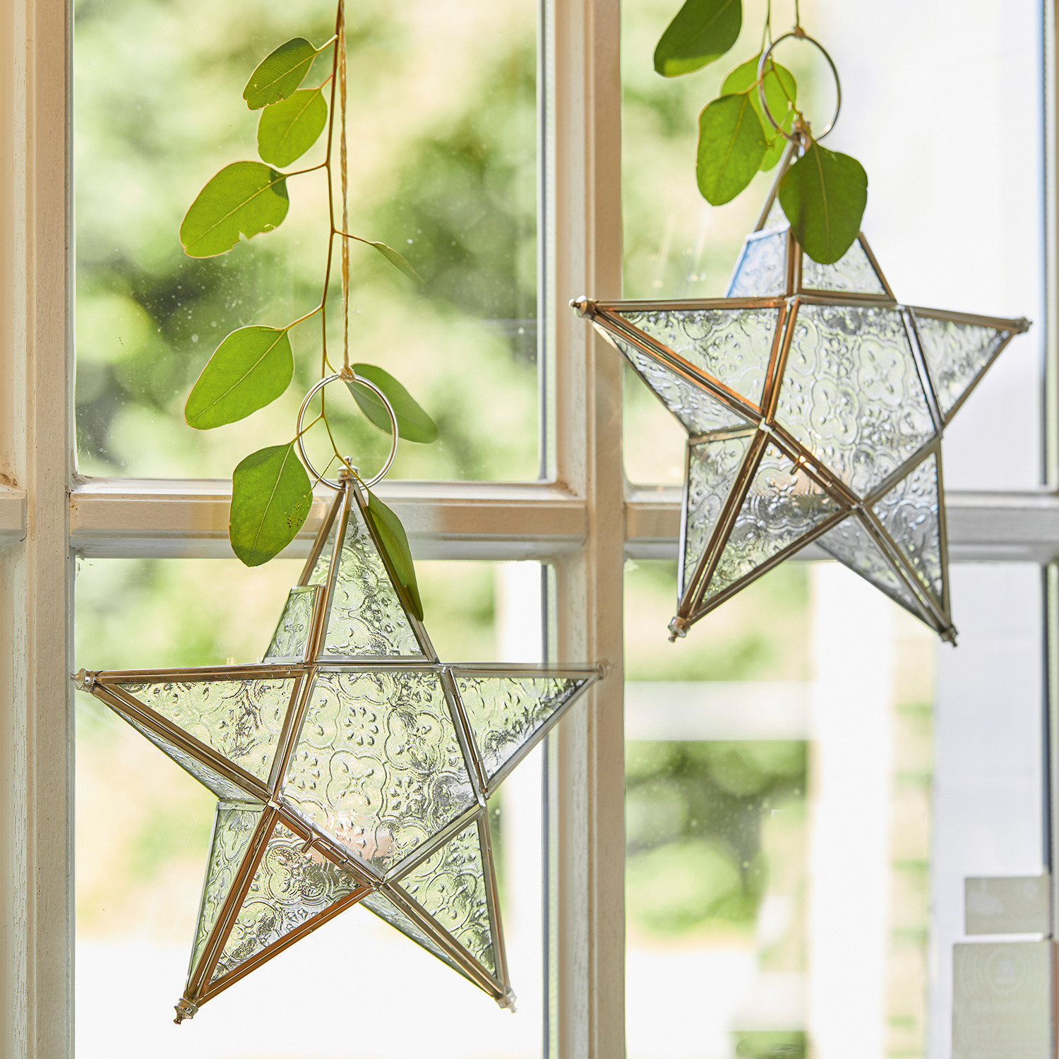 Lot de 2 photophores Kian Étoiles décoratives en verre avec cadre métallique suspendues à une fenêtre avec feuilles vertes