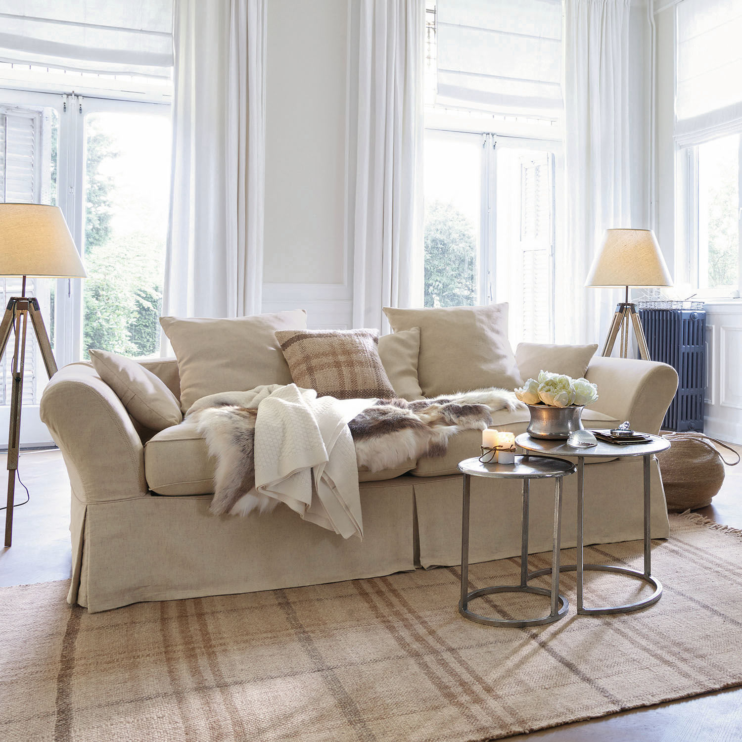 Beige stoffen bank met kussens en bontdeken op lichte vloerkleed in lichte woonkamer met staande lampen