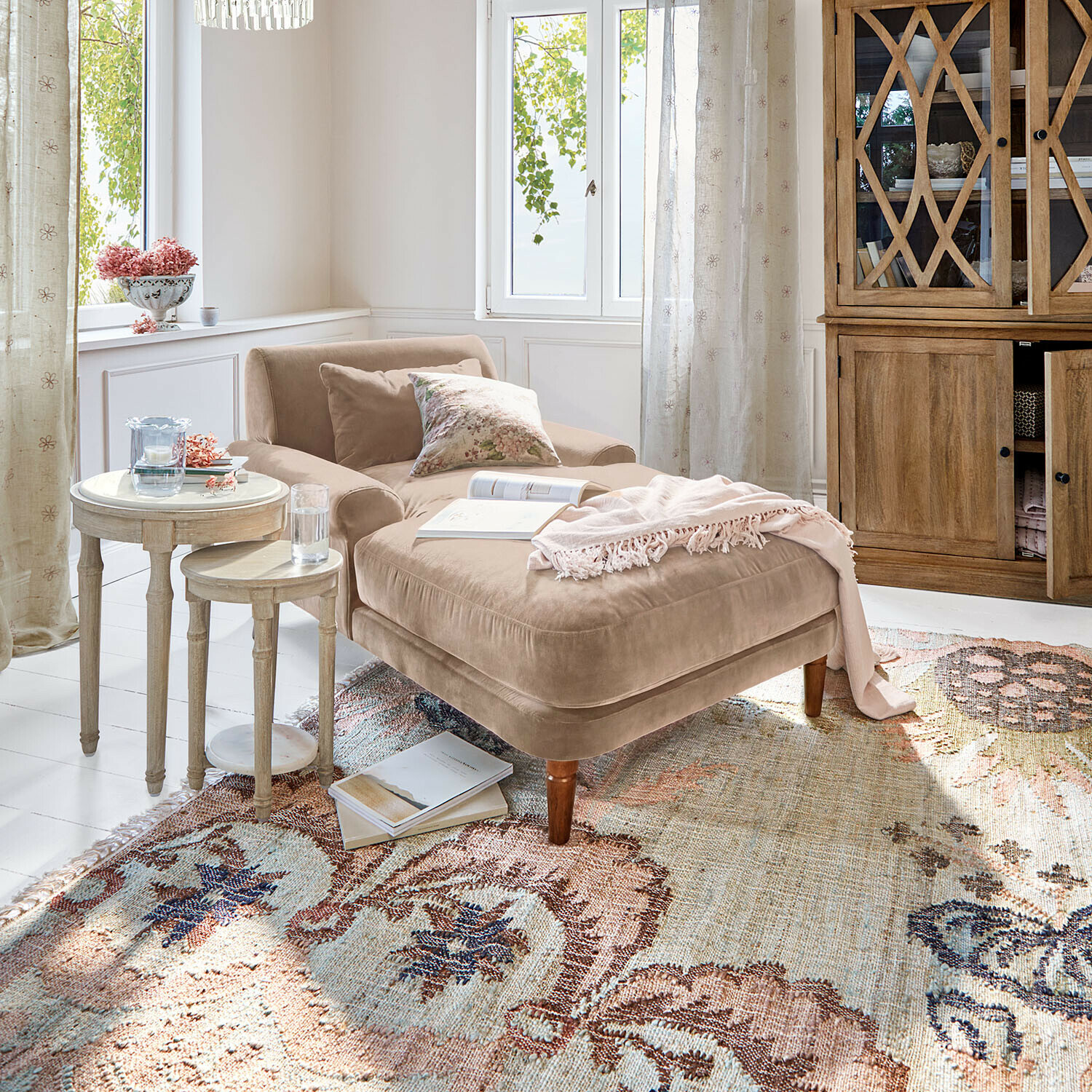 Chaise longue Jouvrelle Beige fluwelen chaise longue met kussens en roze deken op een patroon vloerkleed in lichte woonkamer