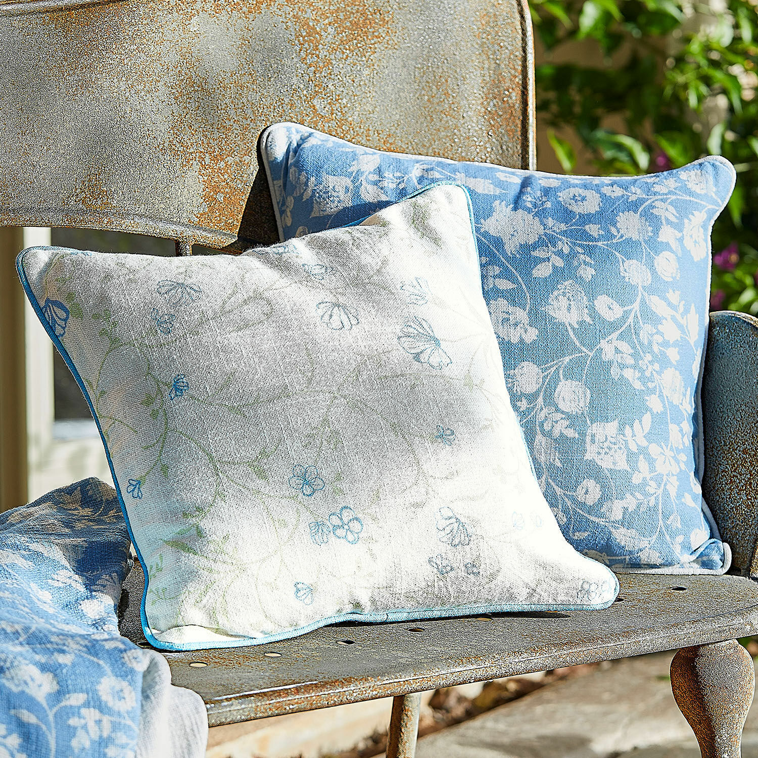 Lot de 2 housses de coussins Corvelique Deux coussins décoratifs en coton bleu et blanc à motifs floraux posés sur un banc en métal rouillé