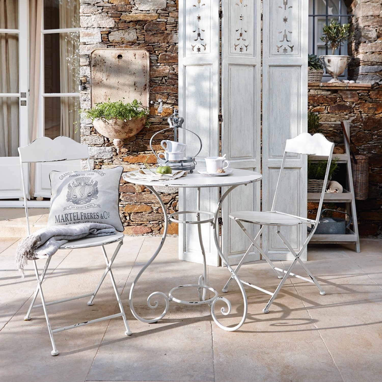 Tuinset Iteuil Witte metalen bistrotafel met twee klapstoelen in vintage stijl op terras met stenen muur