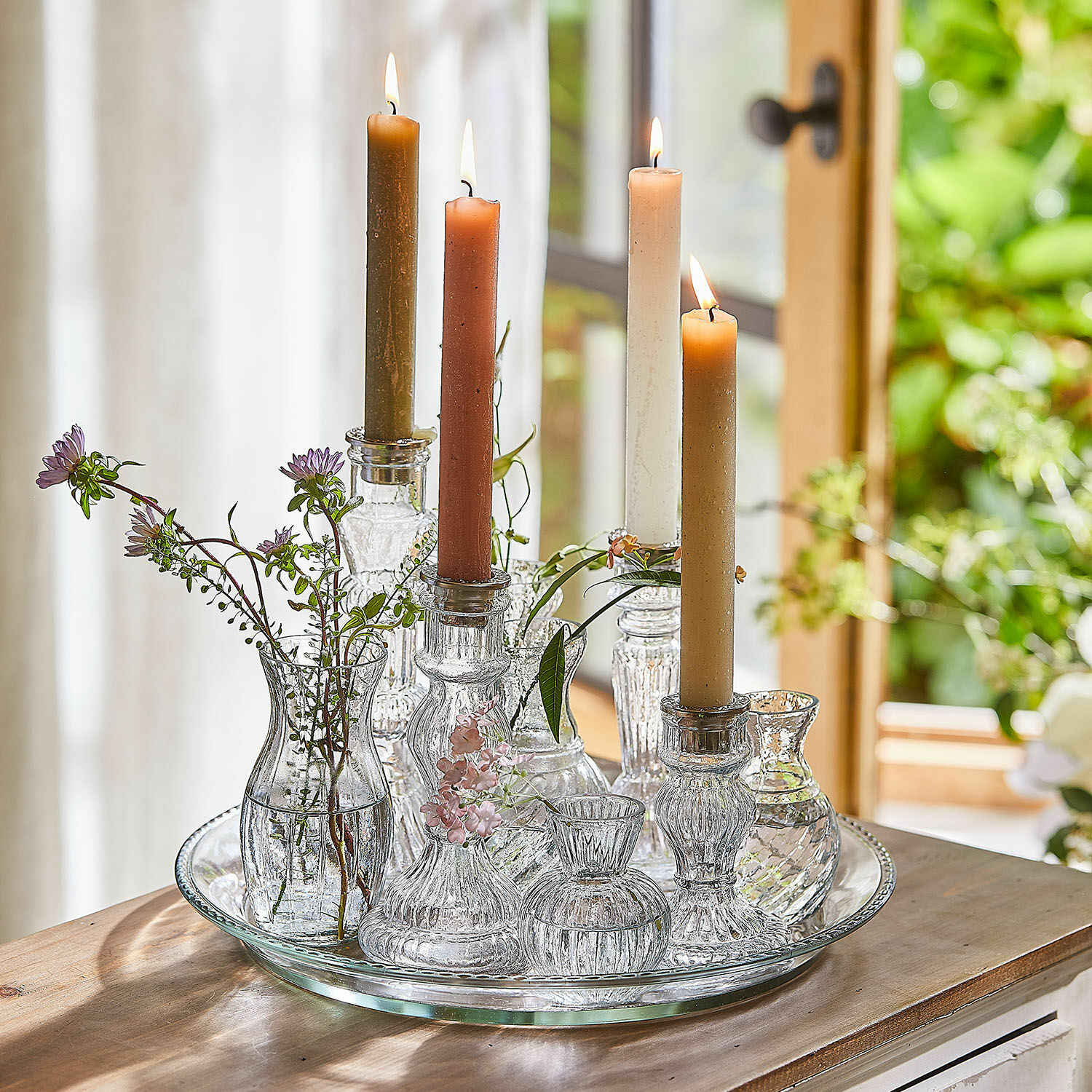 Dienblad met kaarsenhouders en vazen Hilevor Glazen kandelaren en vaasjes met gekleurde kaarsen en bloemen op een glazen dienblad op houten tafel