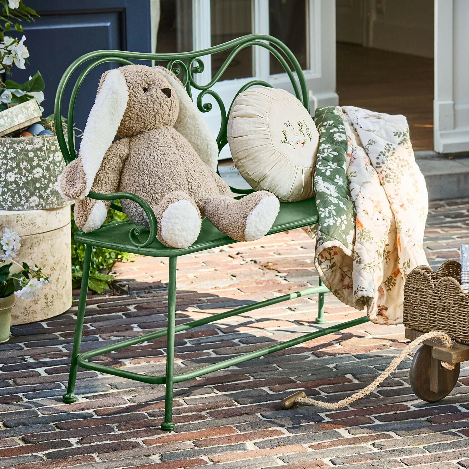 Bankje Fernwick Groene metalen bank met beige pluchen konijn en bloemen deken op een bakstenen terras