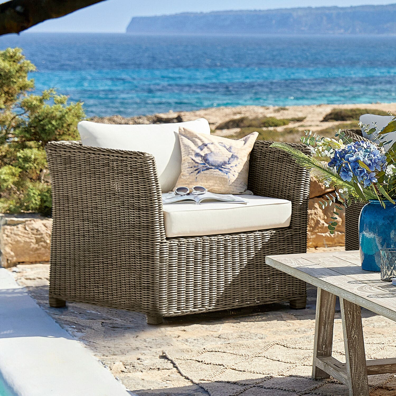 Fauteuil Graves Fauteuil de jardin en rotin gris avec coussins blancs devant la mer et des rochers