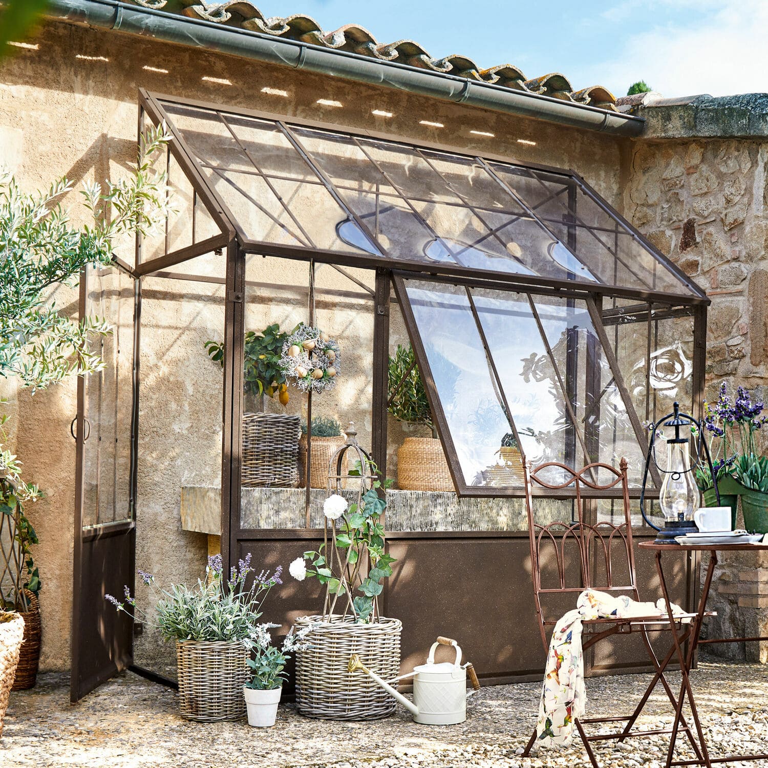 Plantenkas Essone Tuinserre met bruine metalen frame, planten en tuinmeubilair in zonnige buitenruimte