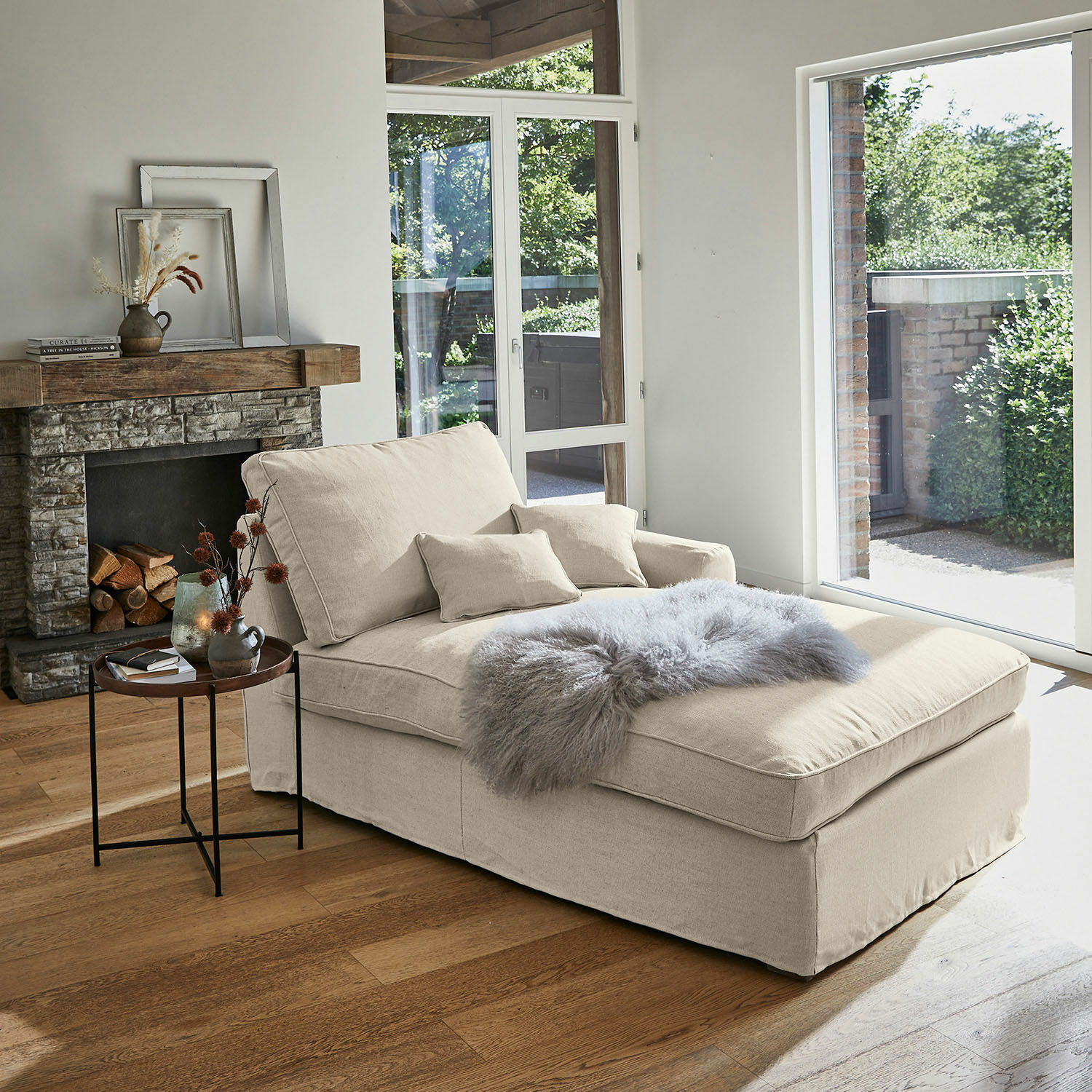 Beige stoffen chaise longue met kussens en grijze bontdeken in lichte woonkamer met open haard
