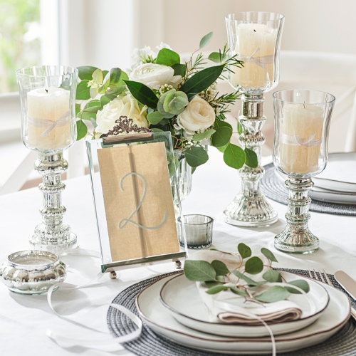 Elegante zilveren kandelaar van glas en metaal met witte kaars op gedekte tafel met bloemen en servies