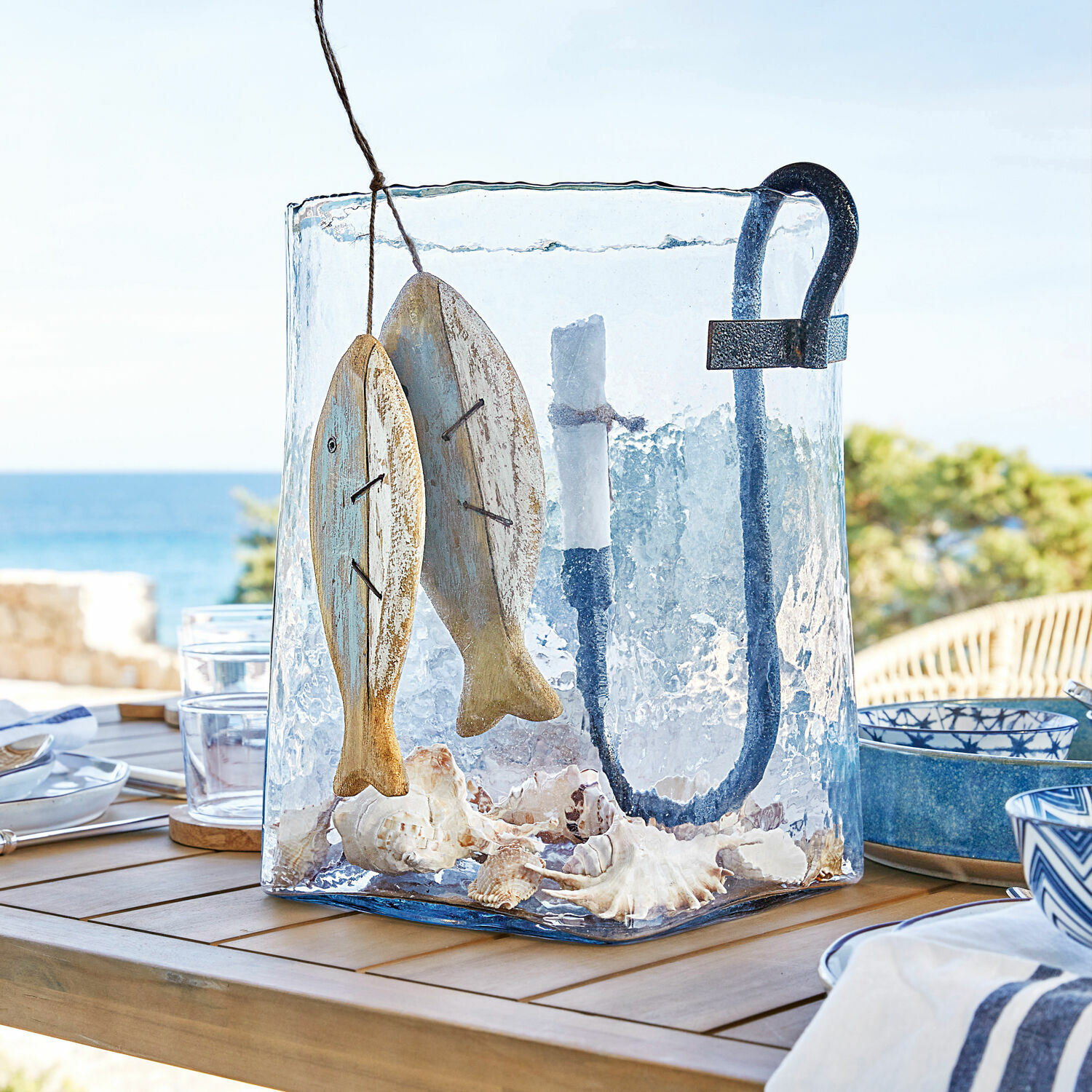 Windlicht Arcene Glazen lantaarn met metalen handvat, houten visdecoratie en schelpen op houten tafel op terras met zeezicht