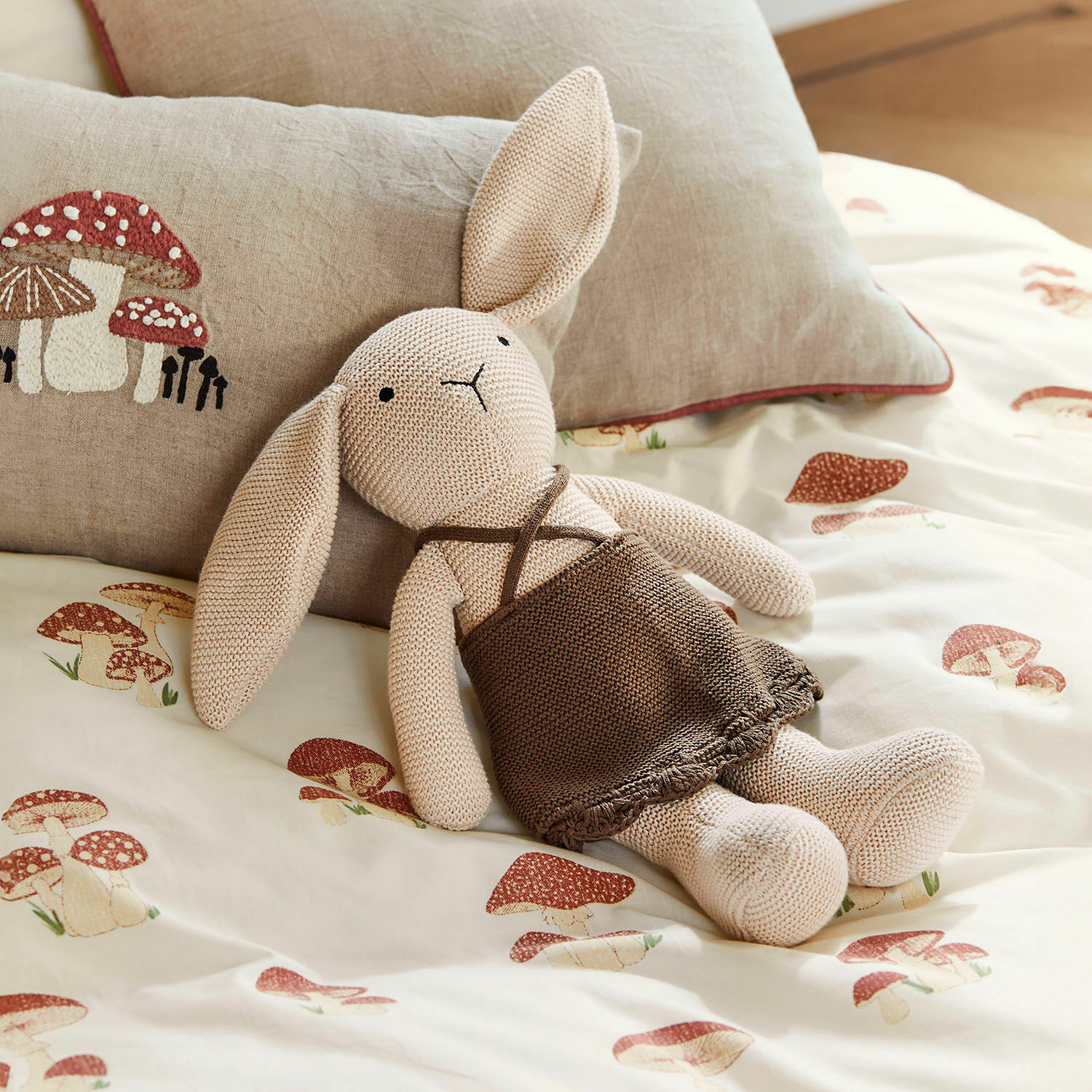 Peluche Flicka Lapin tricoté beige en robe marron sur une literie imprimée de champignons, coussin décoratif champignon