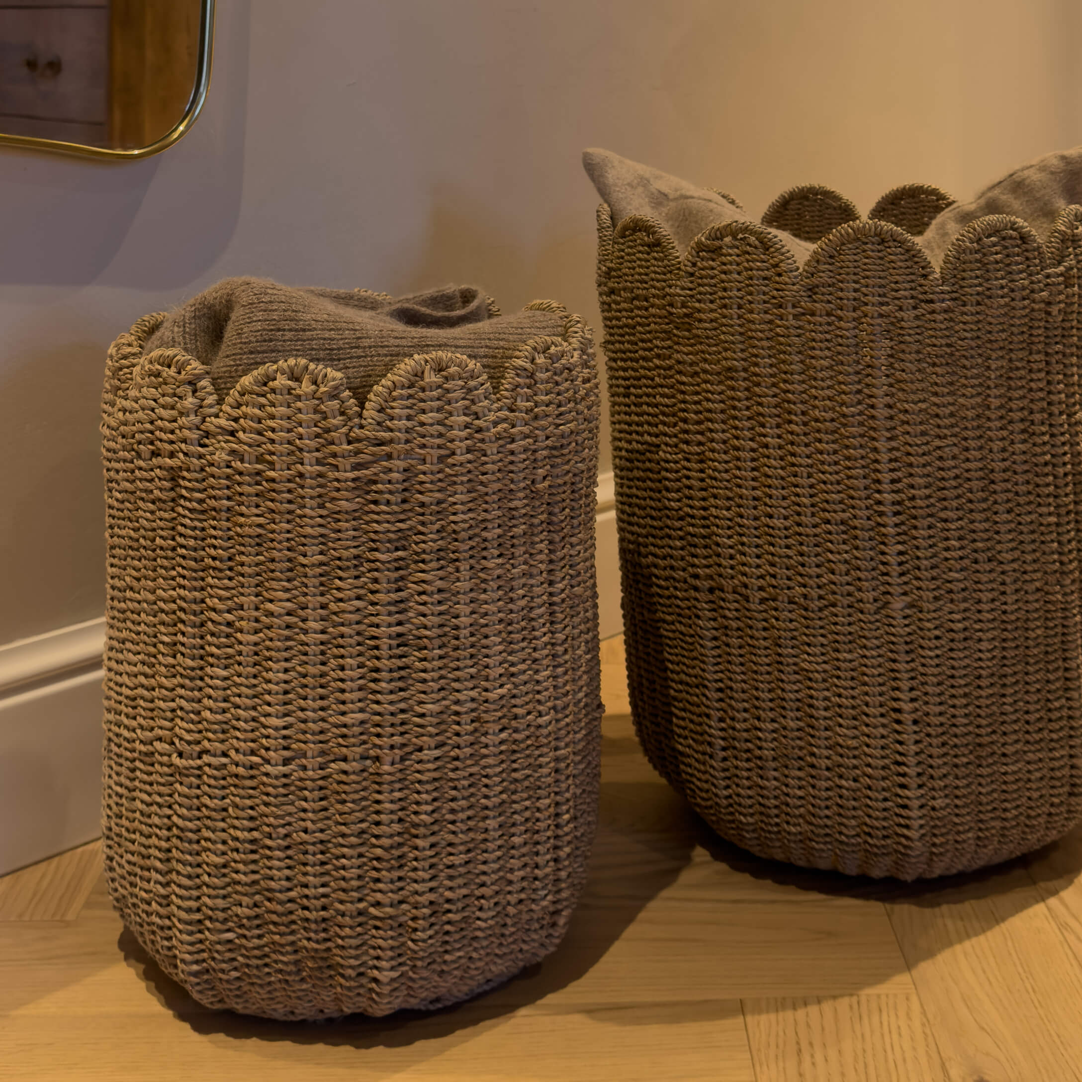 Deux paniers tressés marron avec bord ondulé, remplis de textiles gris sur un sol en bois clair