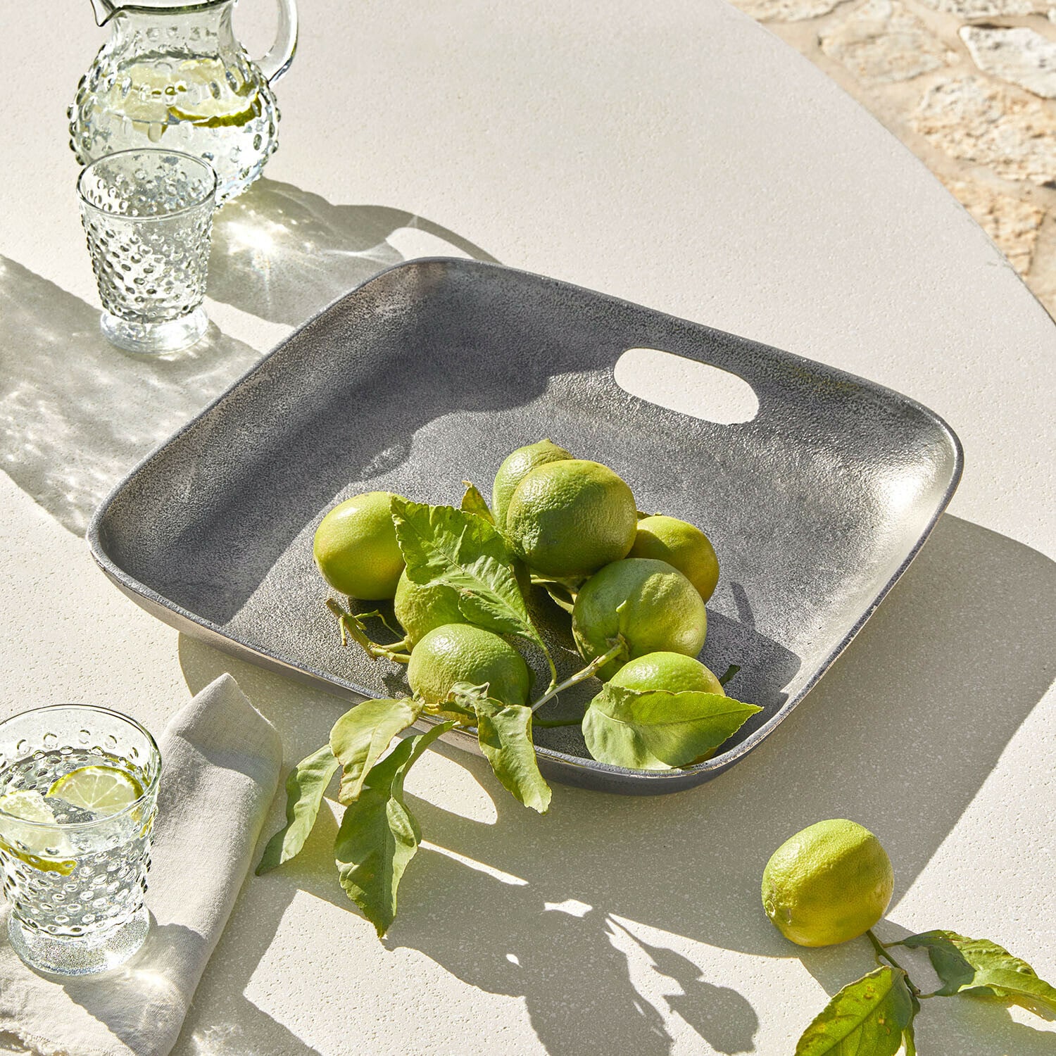 Plateau carré en métal gris avec citrons verts sur une table claire, décoration de table estivale