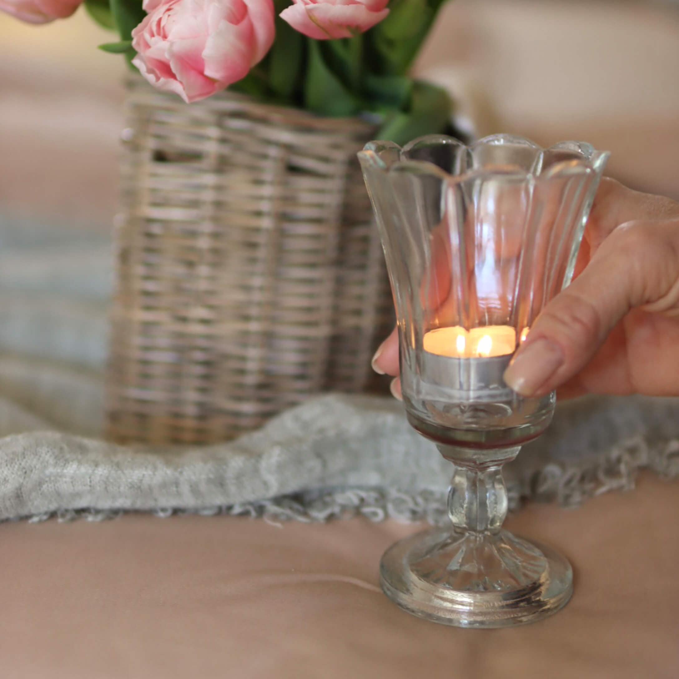 Photophore en verre sur pied avec bougie allumée, vase avec fleurs roses et panier en osier en arrière-plan