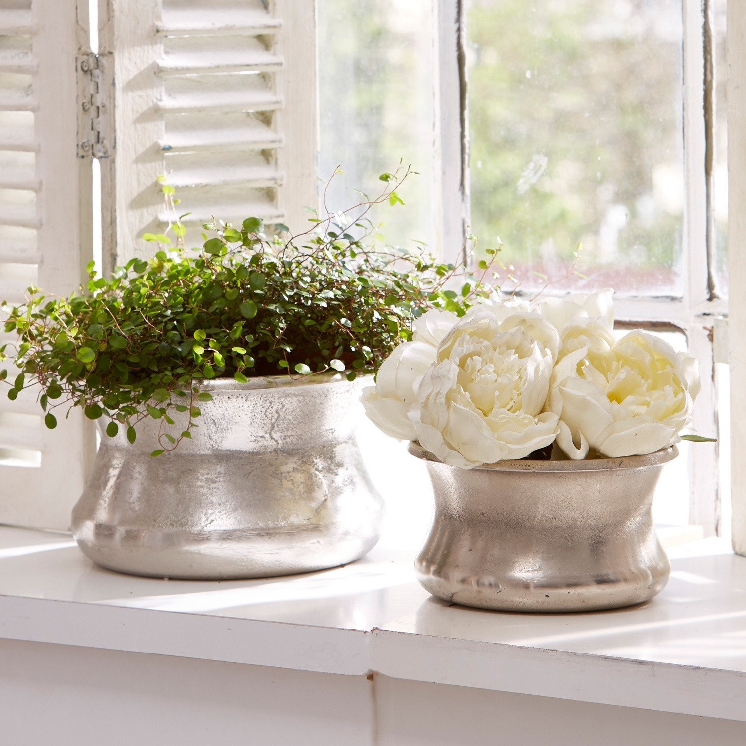 Vaas set van 2 Riona Twee zilverkleurige metalen bloempotten op vensterbank met groene plant en witte decoratieve bloemen