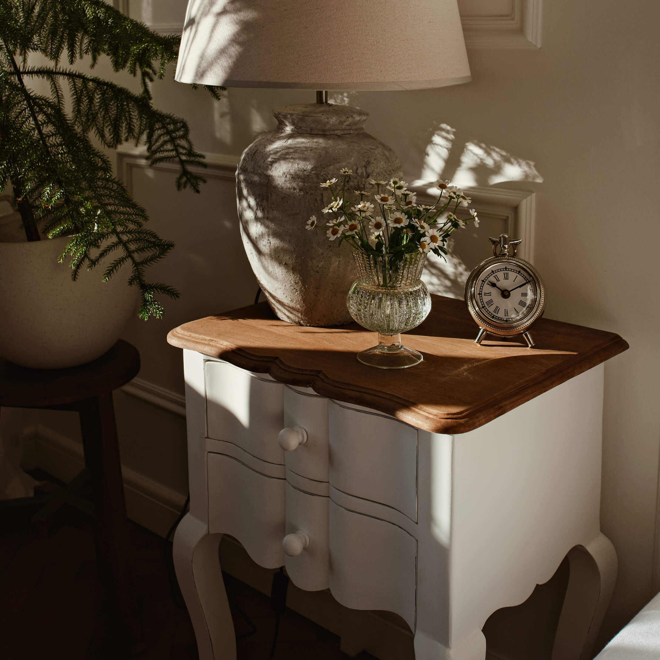 Table de chevet en bois blanc avec plateau marron, lampe, vase en verre avec fleurs et horloge argentée