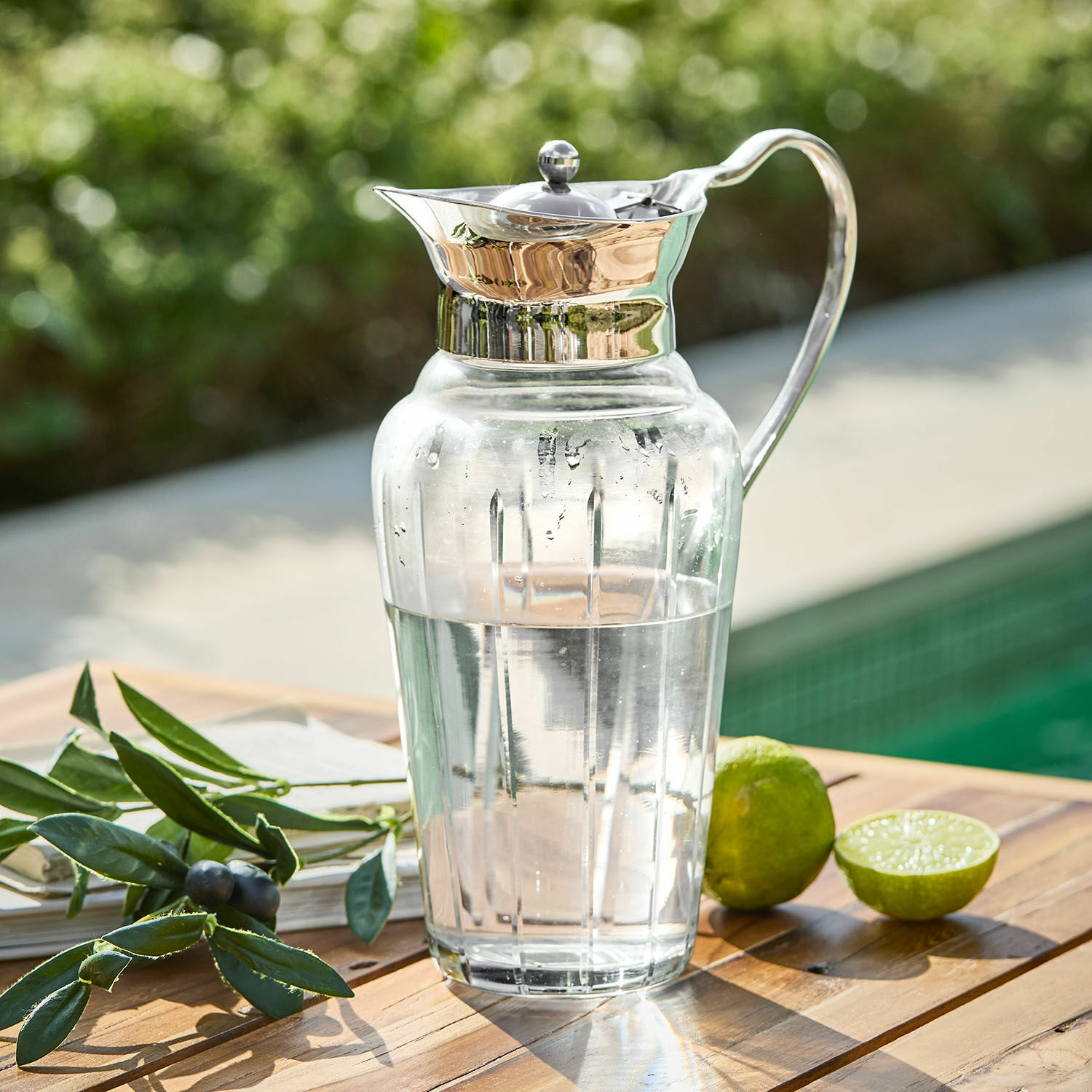 Carafe en verre avec couvercle et poignée en métal remplie d'eau, posée sur une table en bois près d'un citron vert et d'une branche