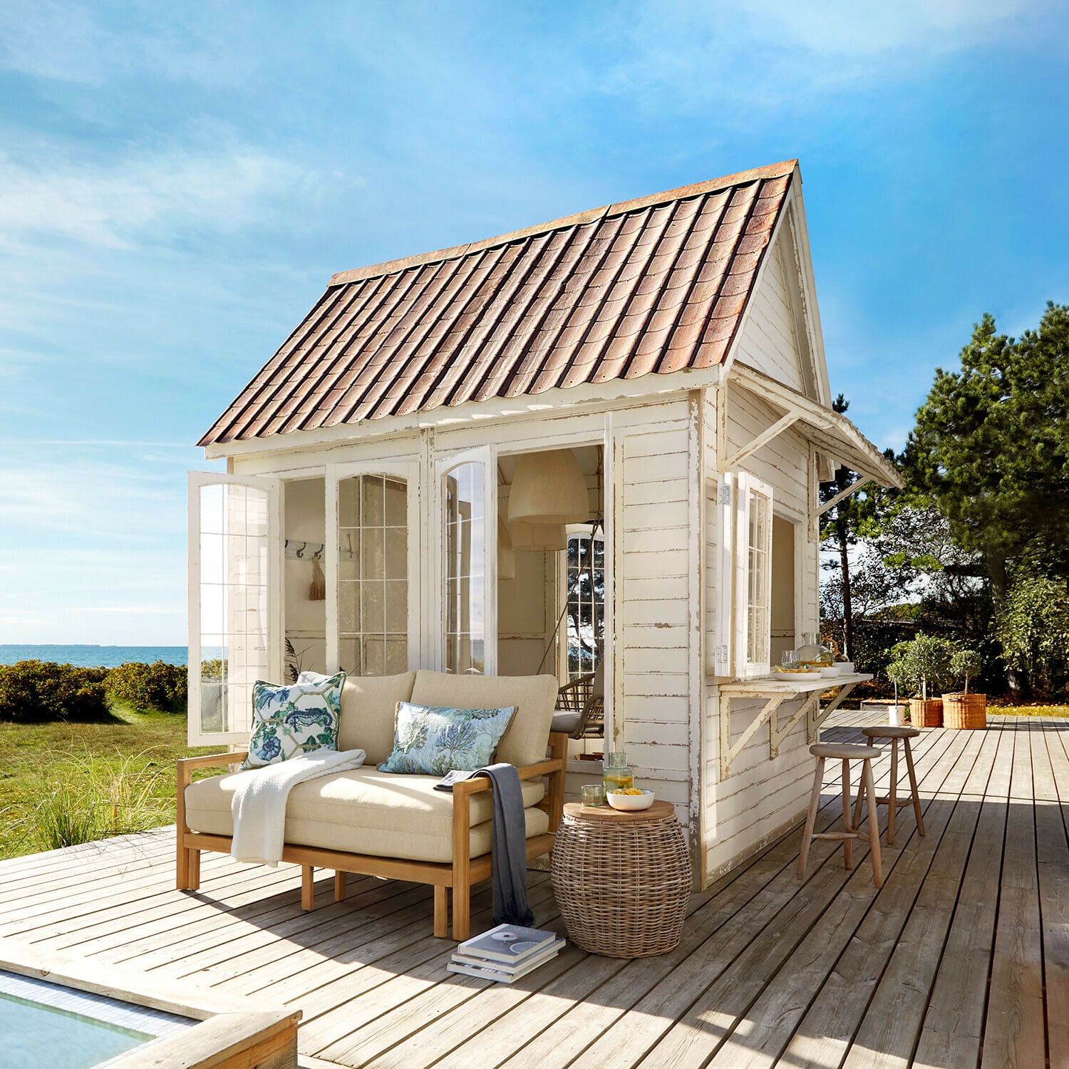Tuinhuisje Stormhavn Witte houten tuinhuis met rood dak en beige bank op houten terras met zeezicht