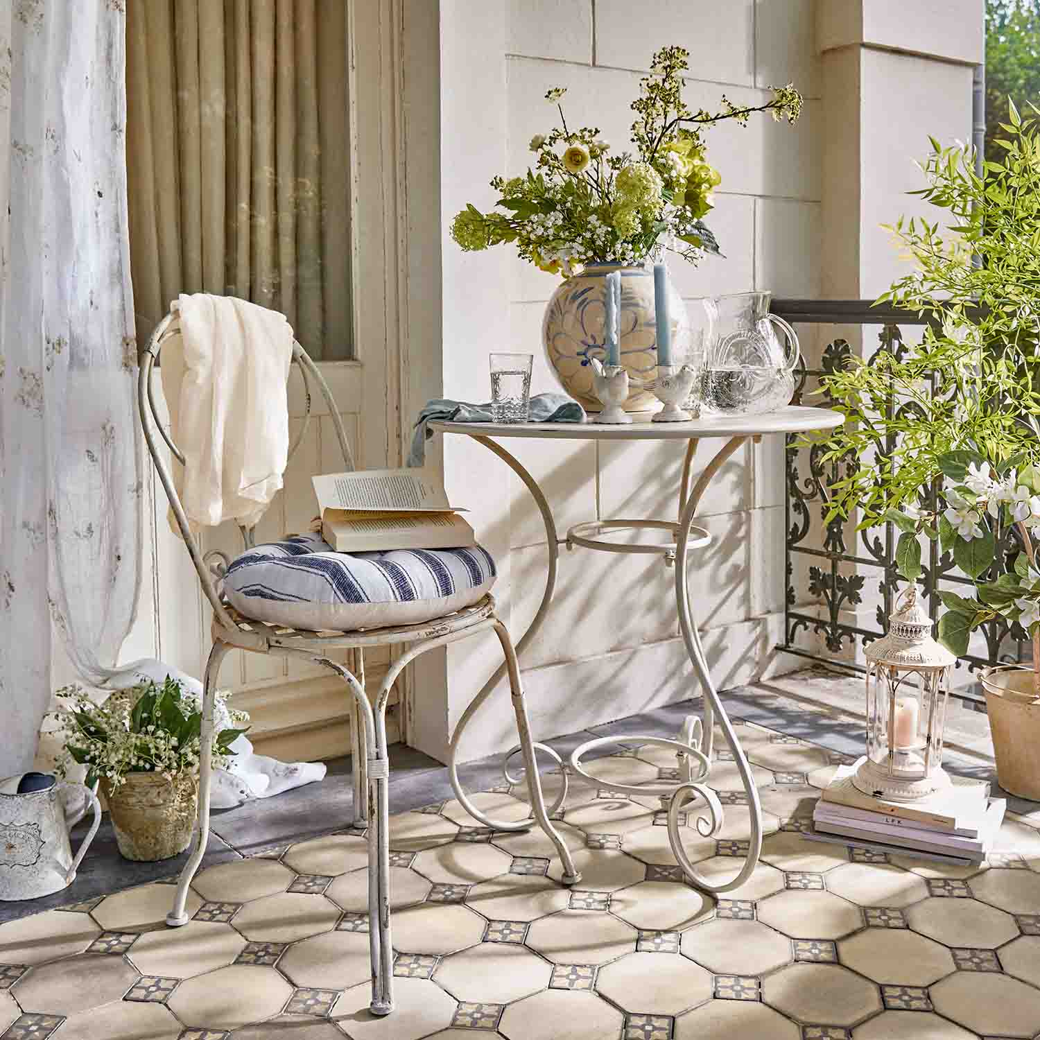 Table et chaise blanches en métal sur terrasse avec coussin rayé, fleurs et lanterne décorative