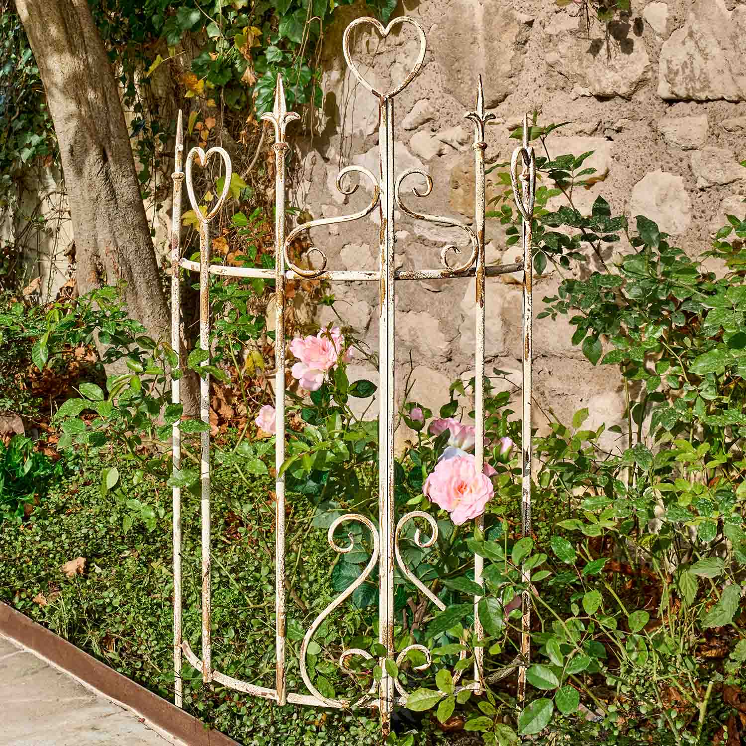 Grille décoratif en métal blanc avec motifs en forme de cœur entourant des roses roses dans un jardin