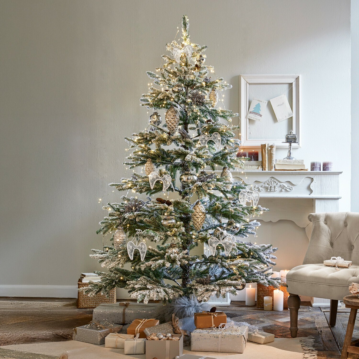 Kerstboom Lokossa Besneeuwde kerstboom met witte en gouden versieringen en cadeaus naast een beige fauteuil in een gezellige woonkamer