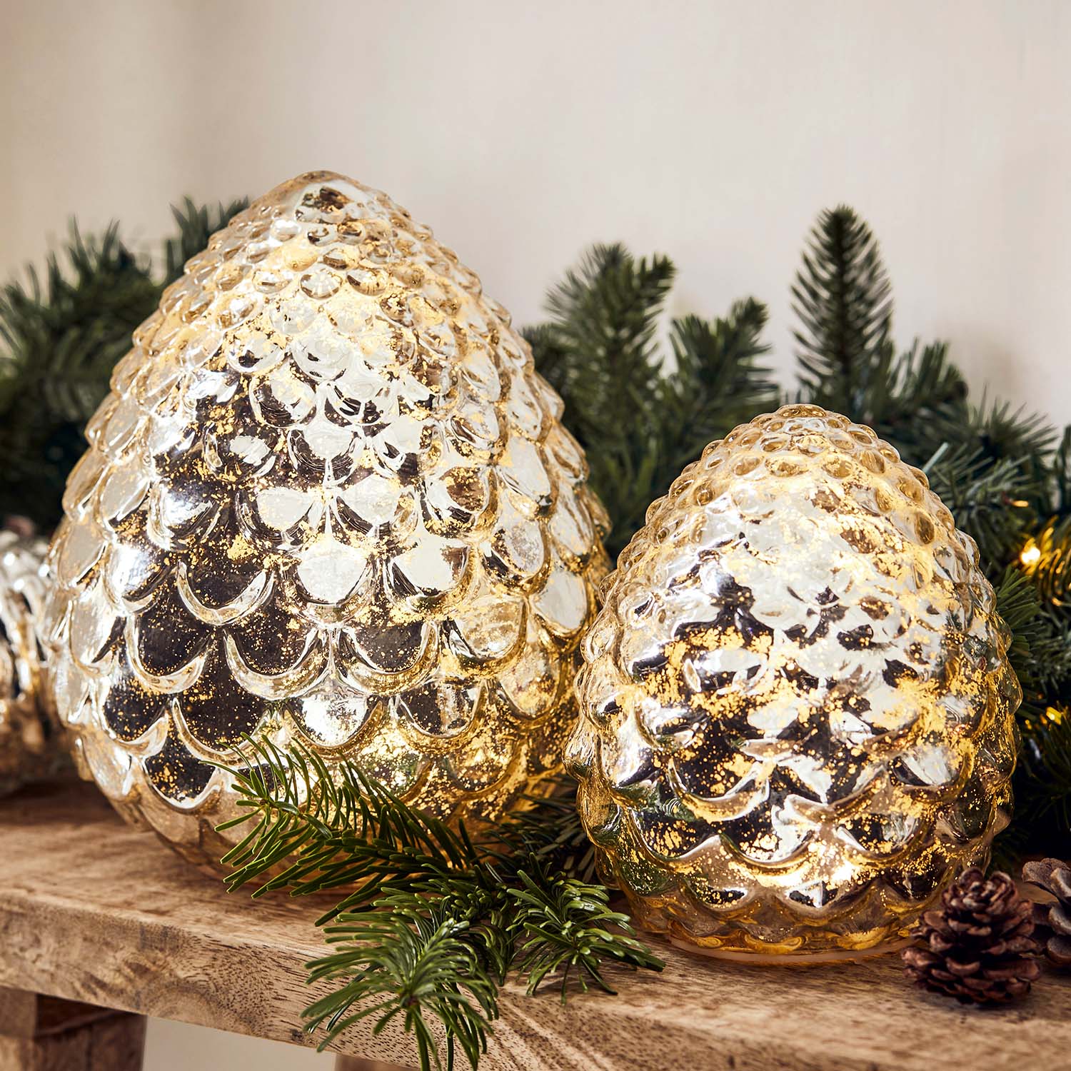 Gouden glazen dennenappeldecoraties op houten plank met groene dennentakken