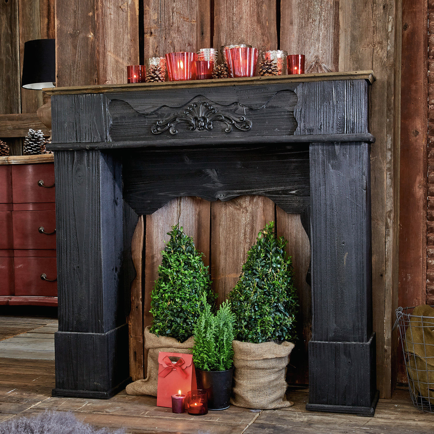 Manteau de cheminée Margaux Cheminée décorative en bois noir avec plantes vertes et bougies rouges dans un intérieur rustique
