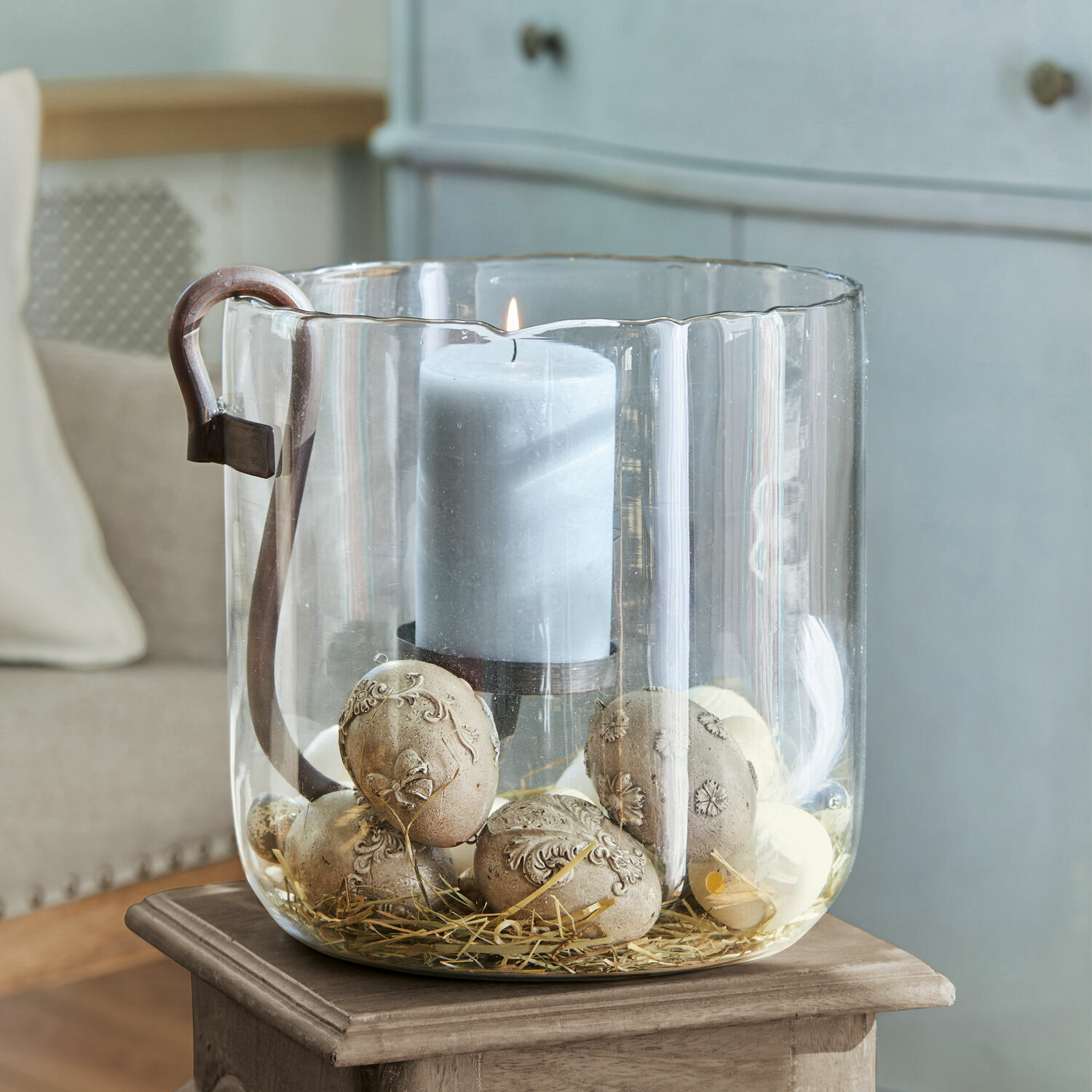 Photophore en verre avec anse, bougie grise et pierres décoratives sur paille pour décoration intérieure