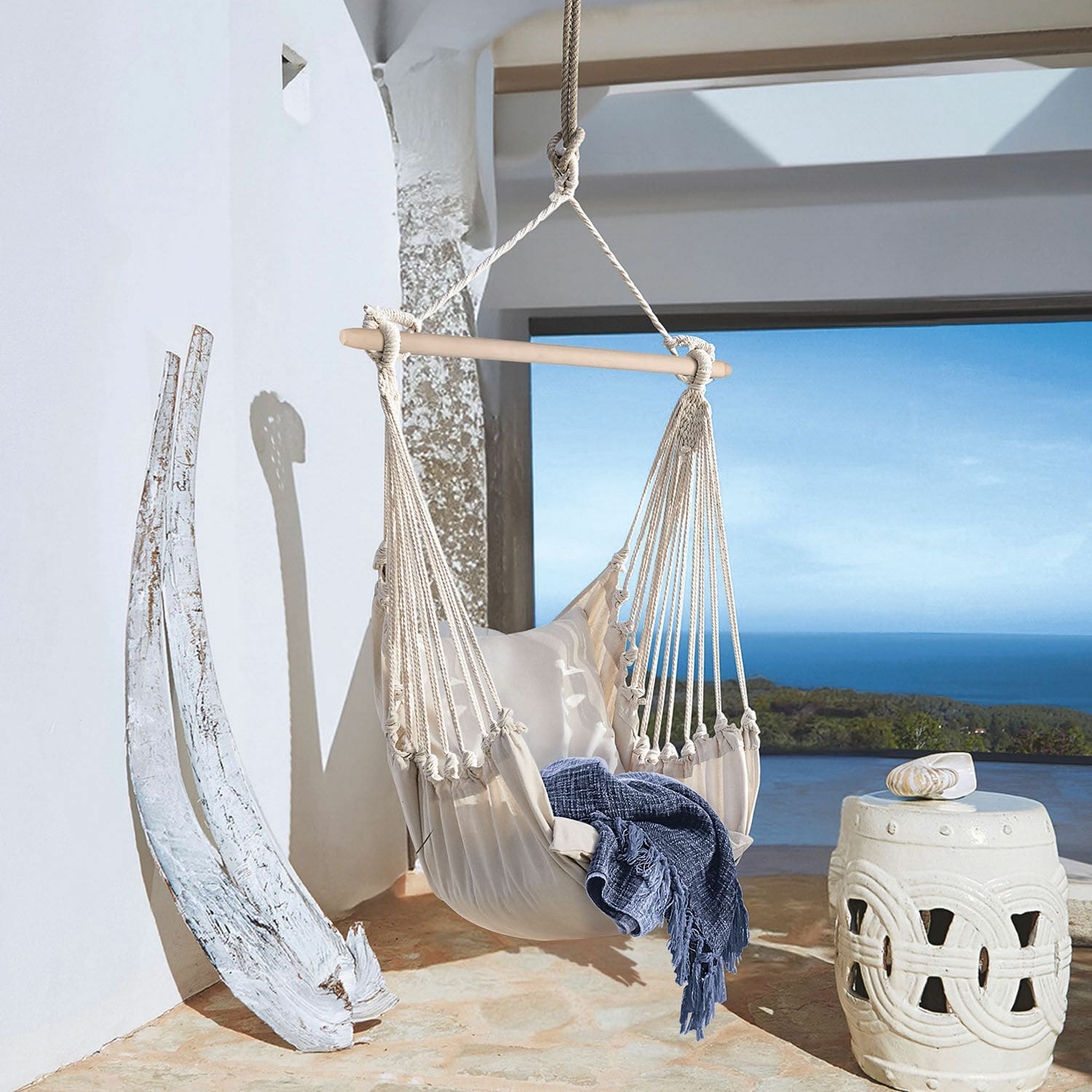 Fauteuil suspendu Leano Hamac beige en coton avec barre en bois et couverture bleue sur terrasse avec vue sur la mer
