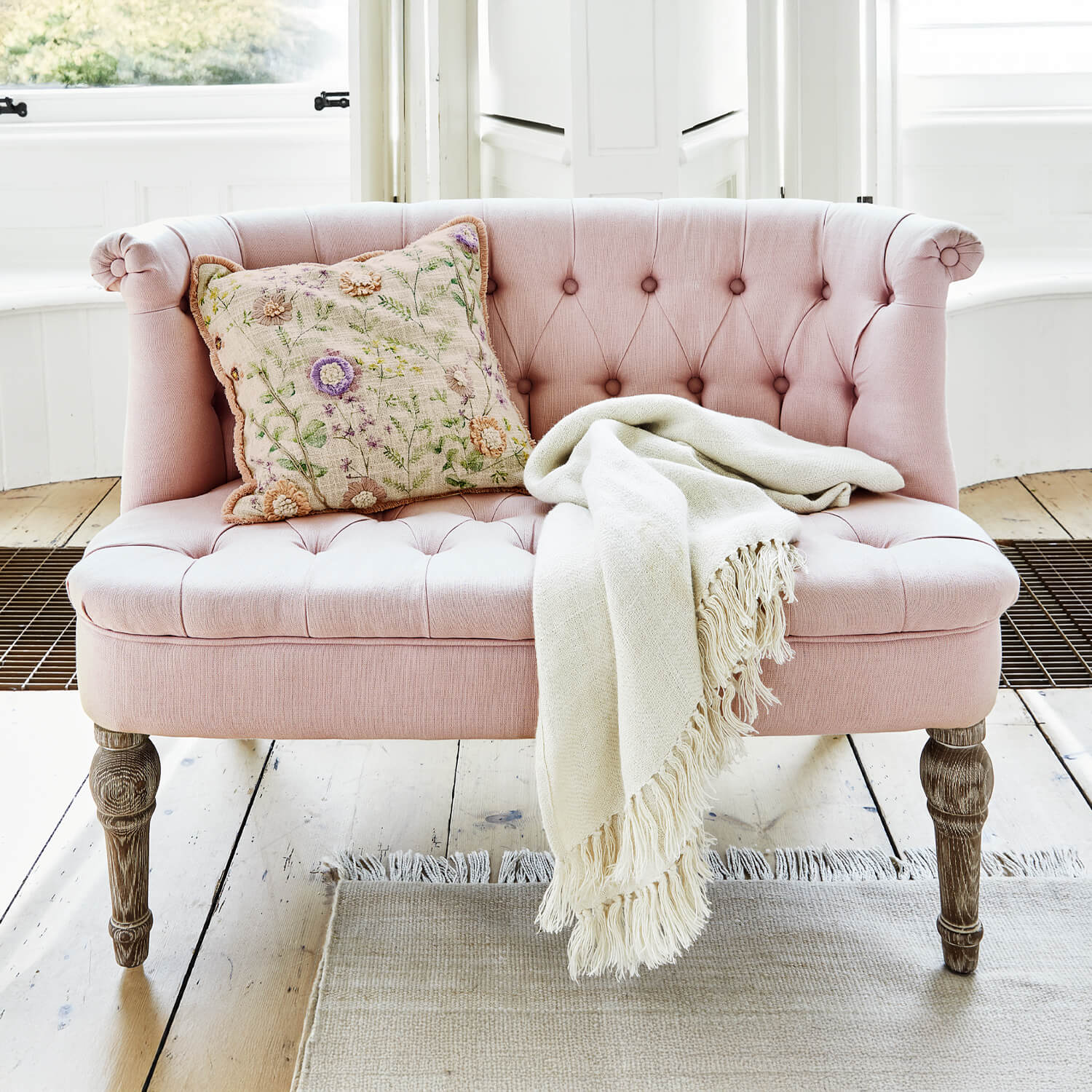 Roze gestoffeerde bank met houten poten, decoratief kussen en crème deken op lichte vloer