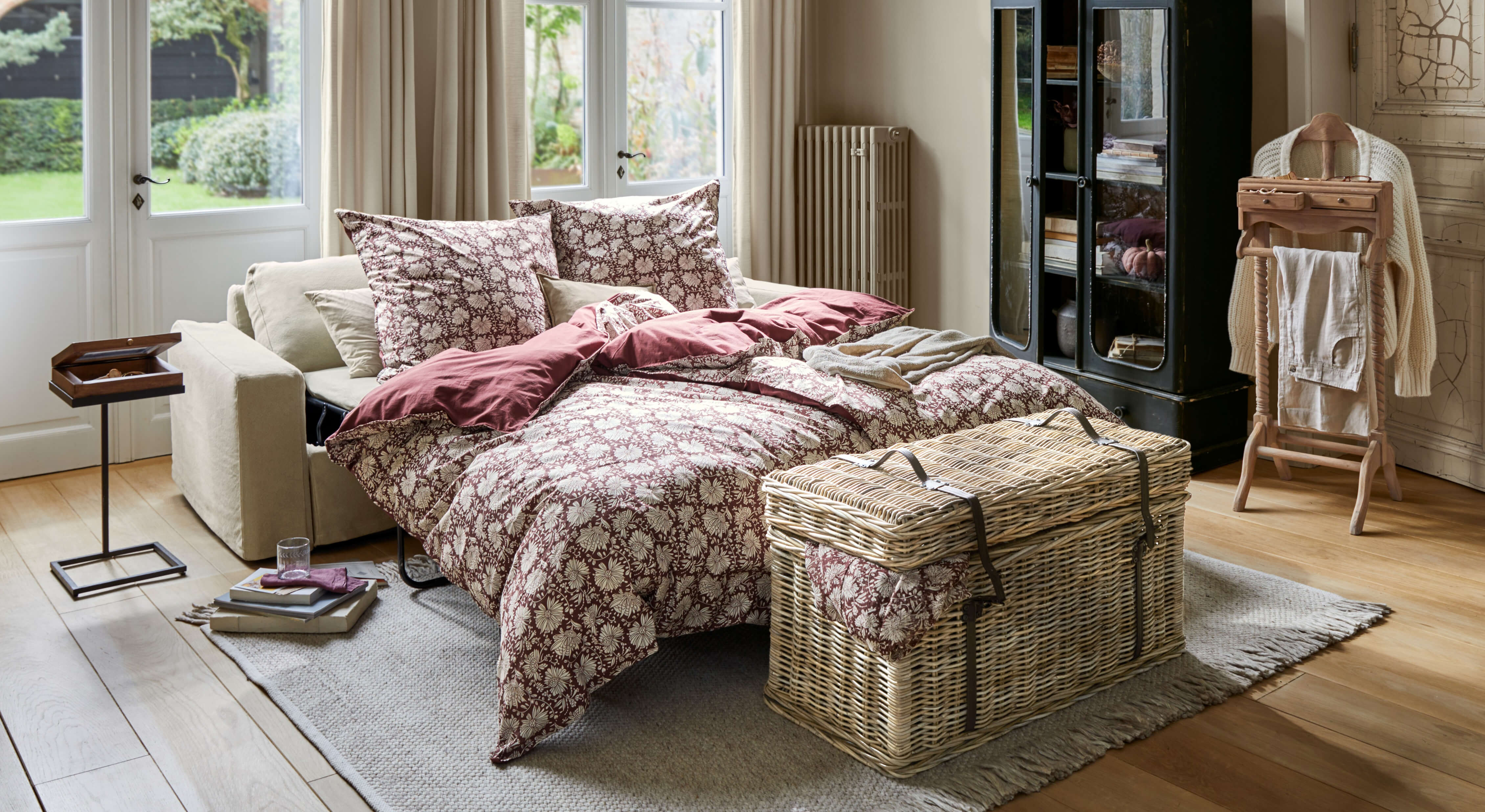 Canapé-lit beige avec literie bordeaux à motif floral dans un salon lumineux au sol en bois