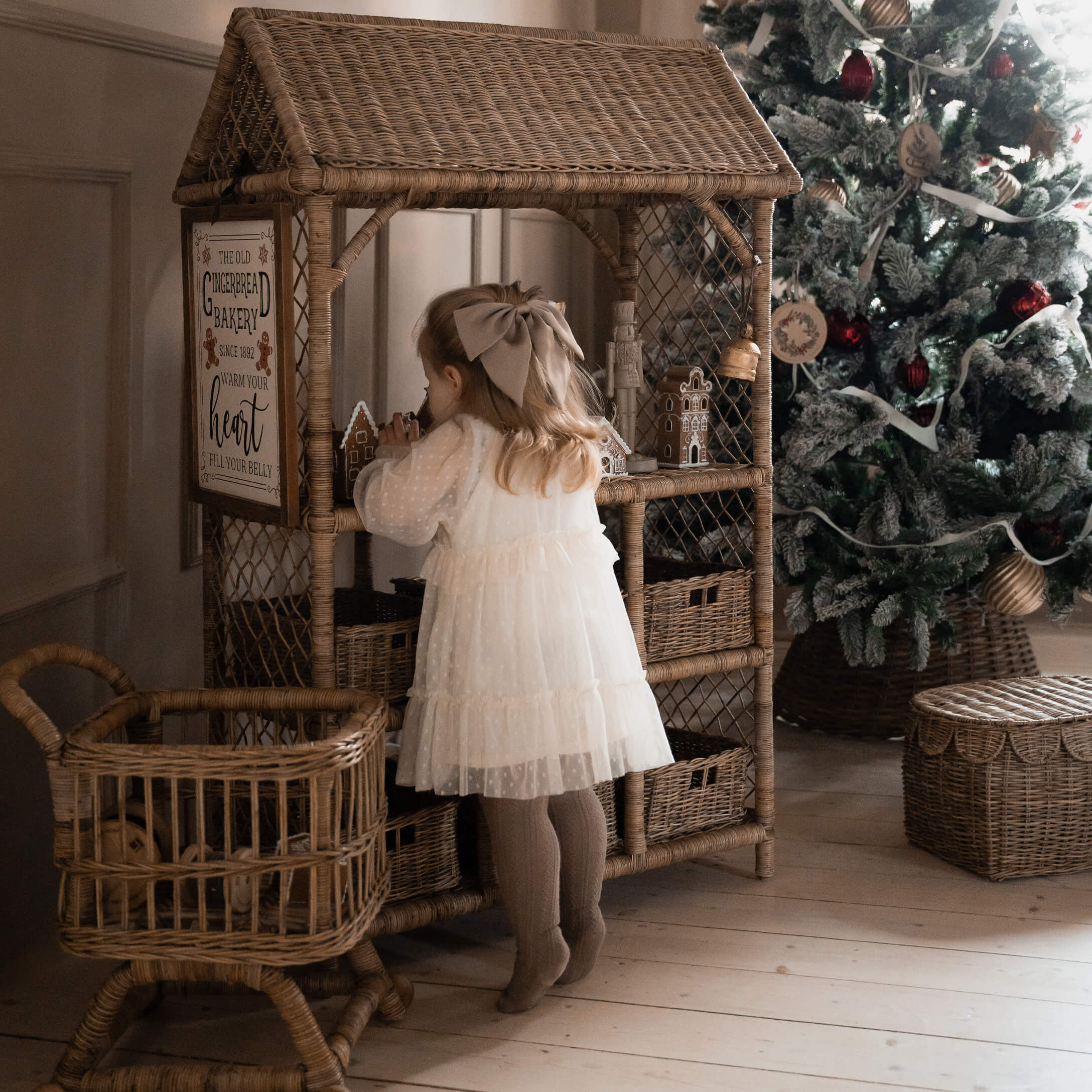 Enfant en robe blanche devant une étagère en osier avec des maisons en pain d'épice près d'un sapin de Noël décoré