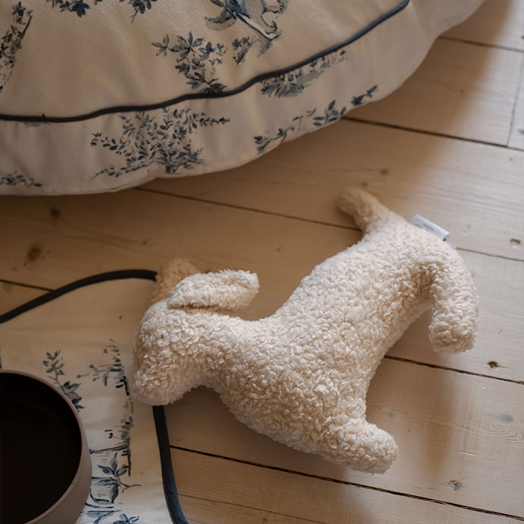 Jouet en peluche douce en forme de chien beige clair posé sur un sol en bois clair