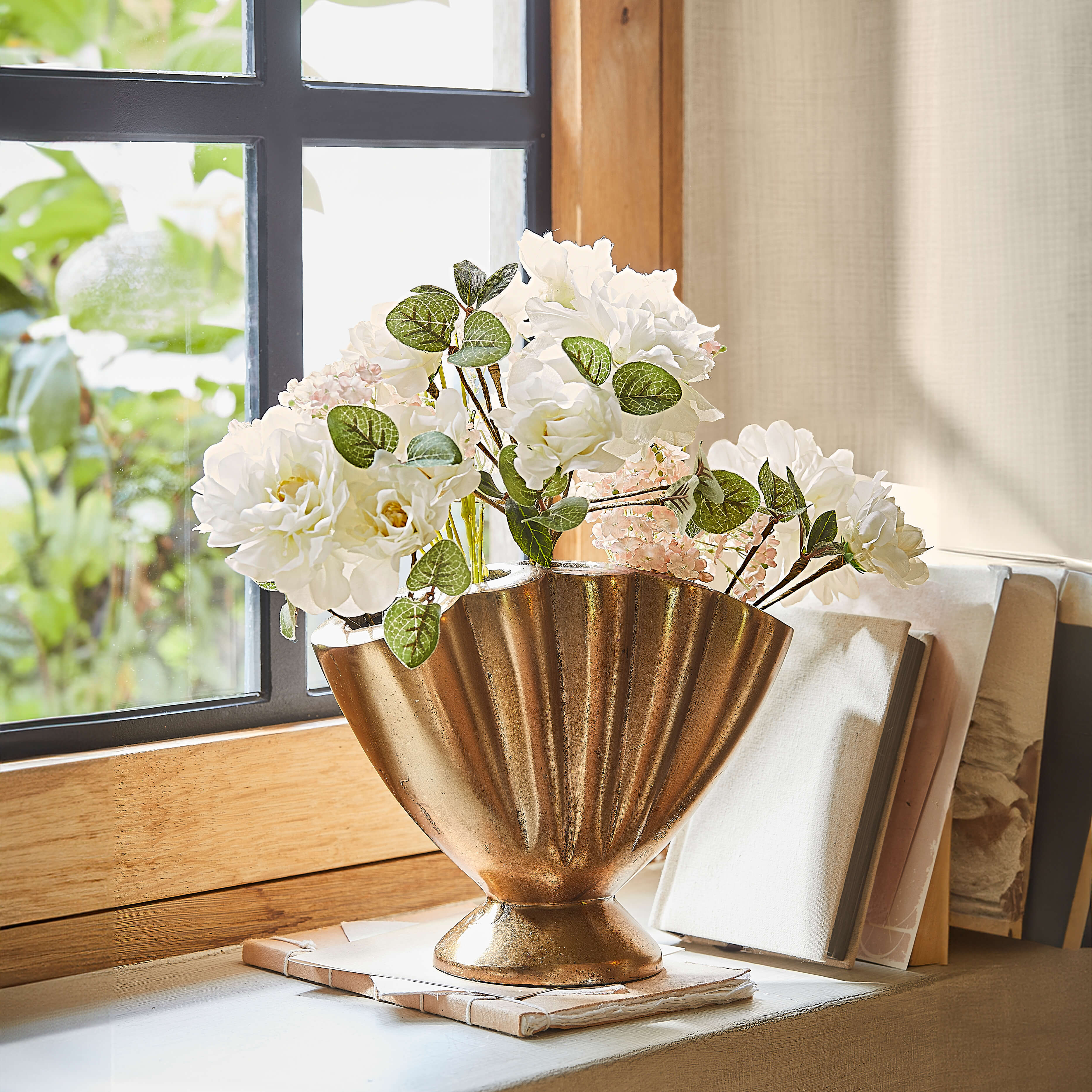 Gouden metalen vaas in waaiervorm met witte bloemen op vensterbank bij daglicht