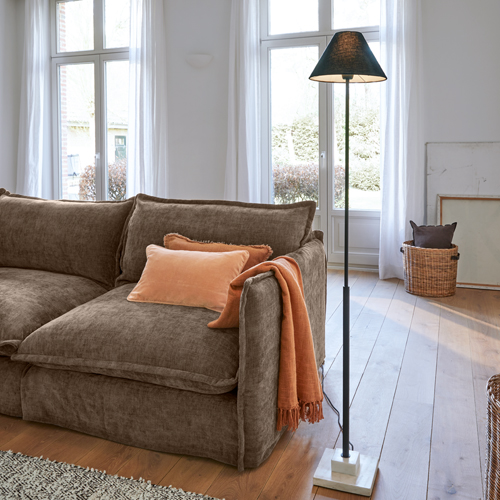 Canapé marron en tissu doux avec coussin orange et plaid, lampe sur pied noire avec base en marbre dans un salon lumineux