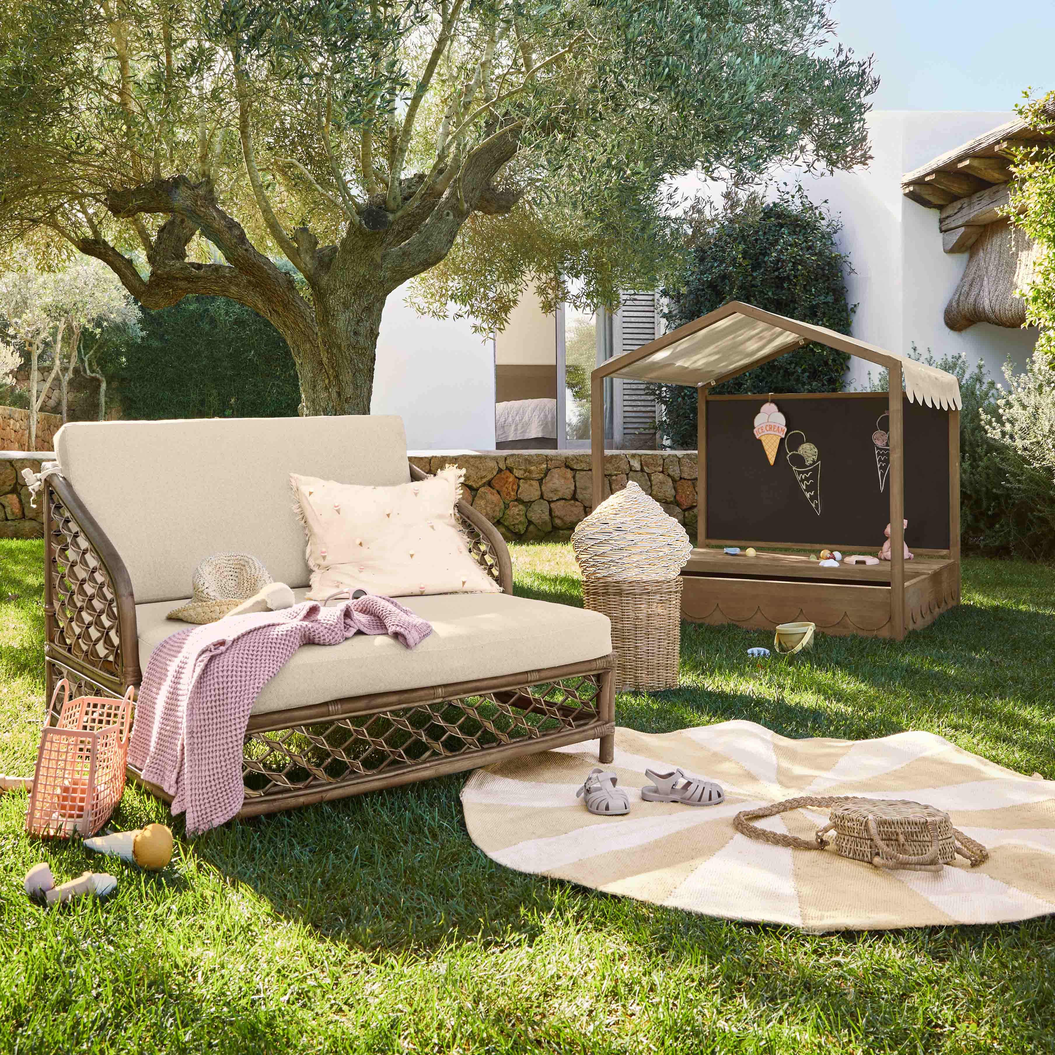 Jardin avec chaise longue beige, coussin et couverture rose sur l'herbe sous un olivier