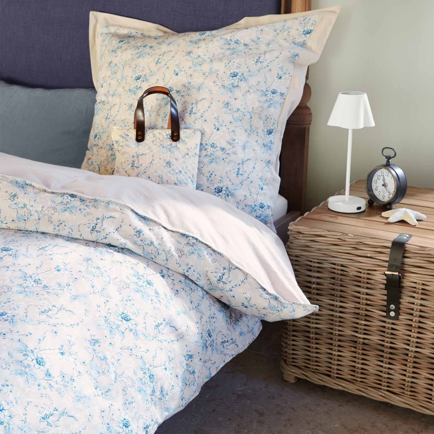 Parure de lit en coton clair avec motif floral bleu sur lit à côté d'une table de chevet en osier