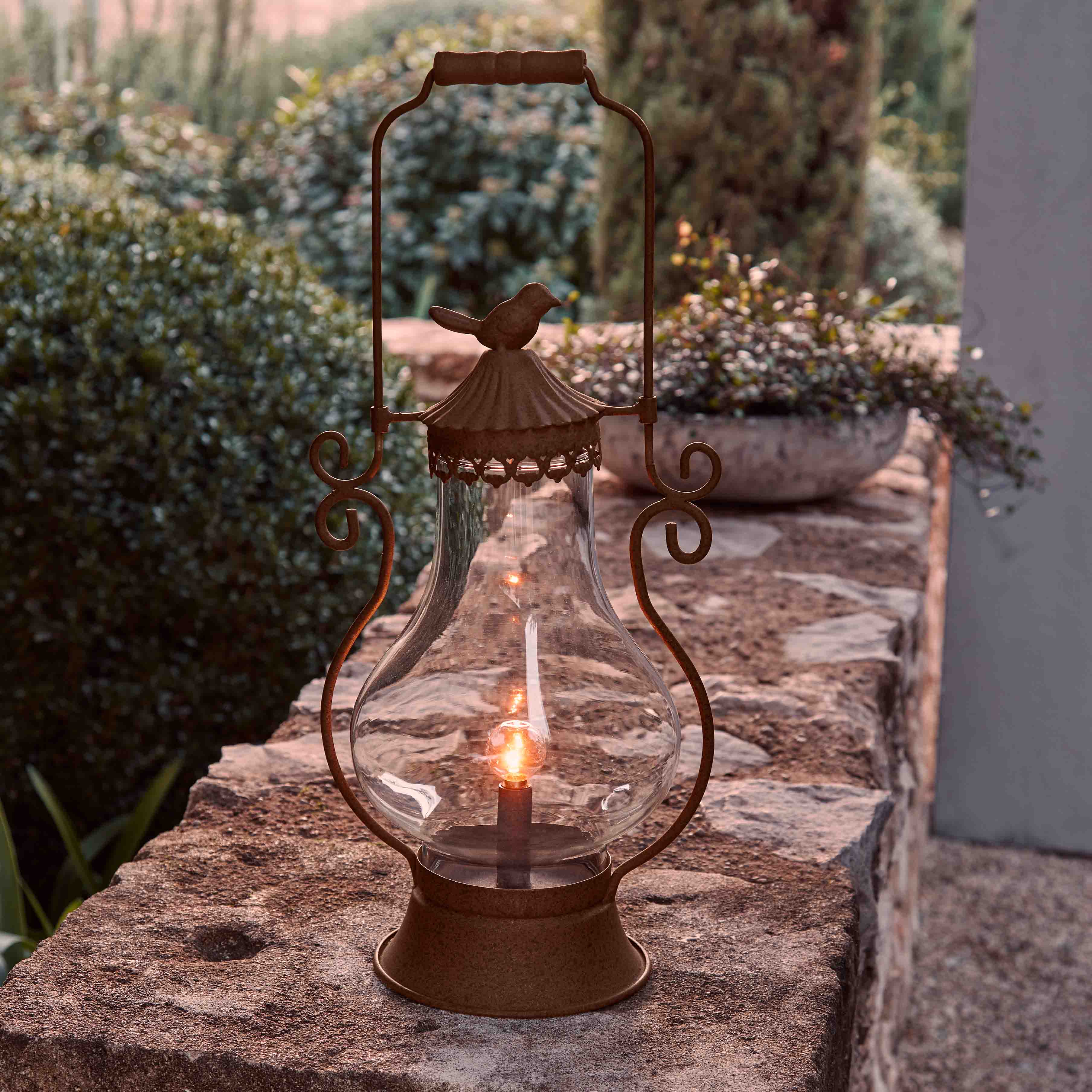 Tuinlantaarn van roestkleurig metaal met helder glas en vogelornament, sfeerverlichting buiten