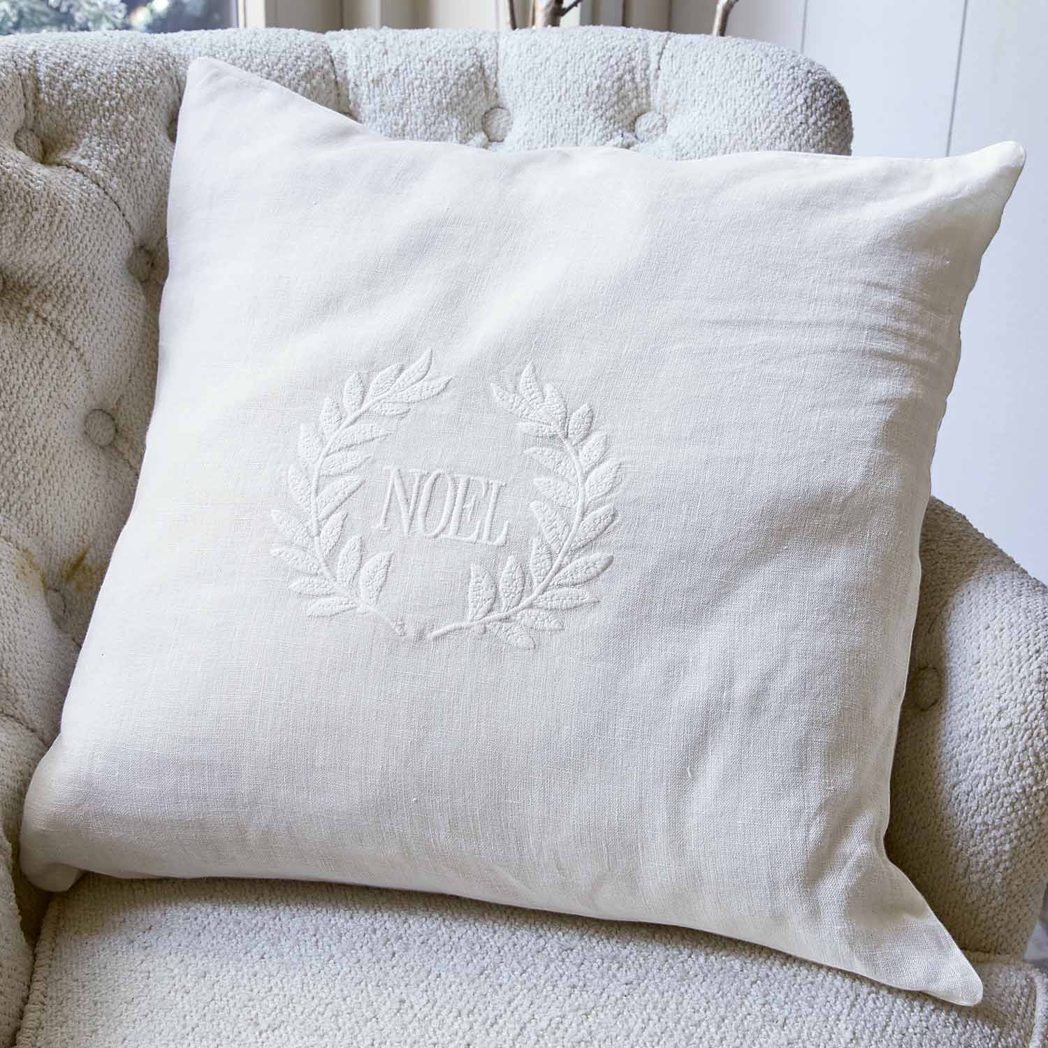 Coussin blanc en lin avec broderie NOEL et couronne décorative sur fauteuil clair, accessoire de Noël