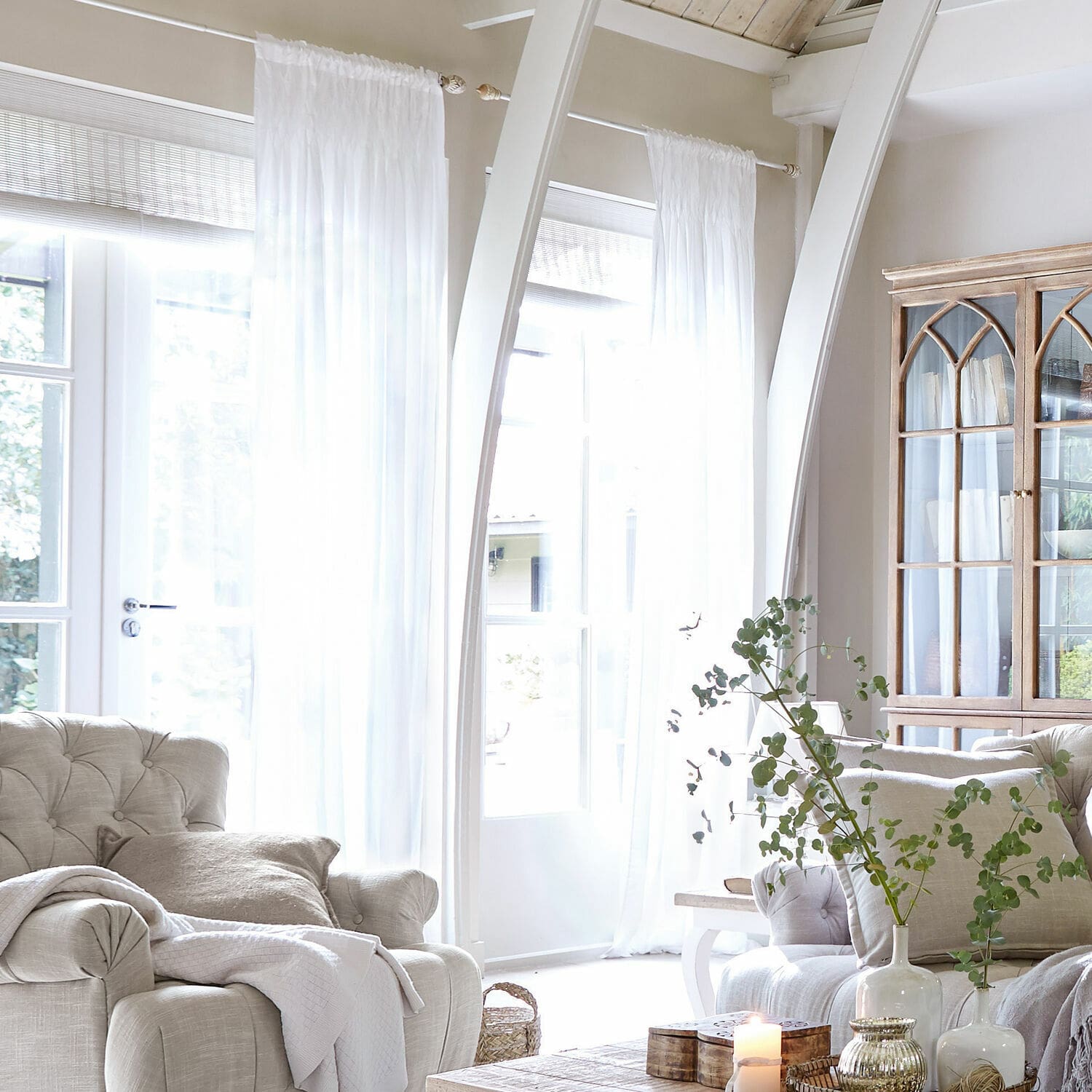 Rideau Otilie Salon lumineux avec fauteuil beige, rideaux blancs et vitrine en bois style vintage