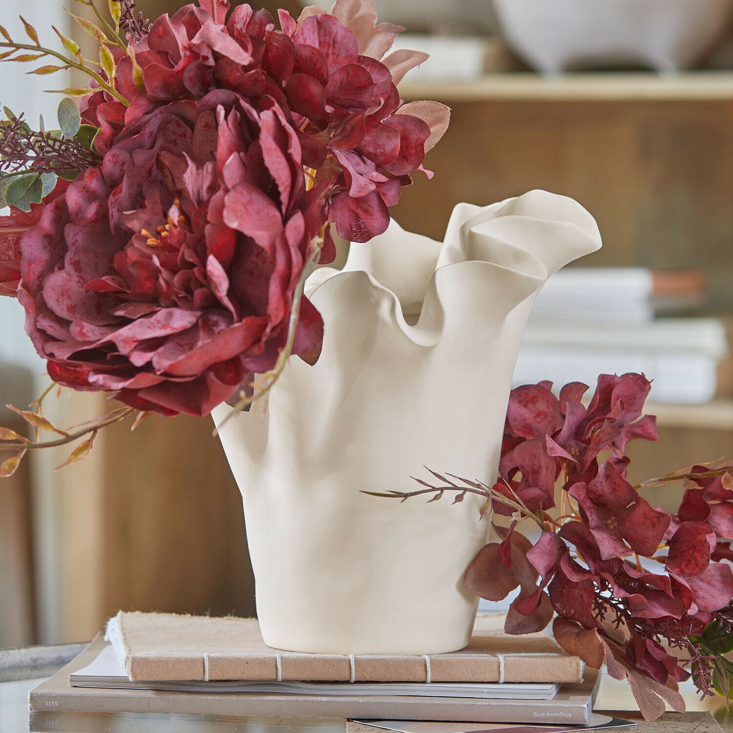 Vase Elvessa Vase en céramique blanche aux bords ondulés avec fleurs artificielles rouges sur table avec livres
