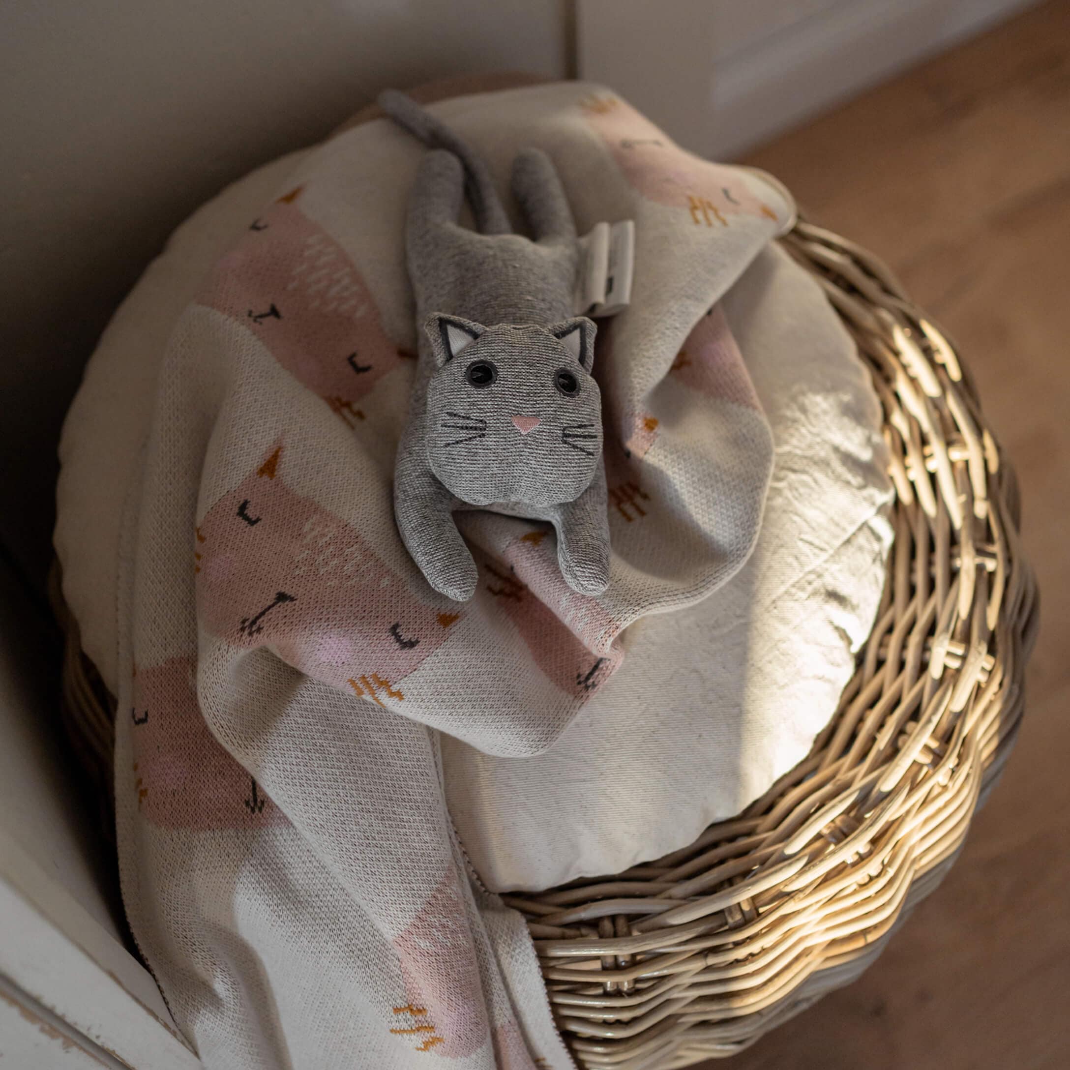 Chat en peluche gris sur une couverture rose clair à motif chat posée sur un panier en osier