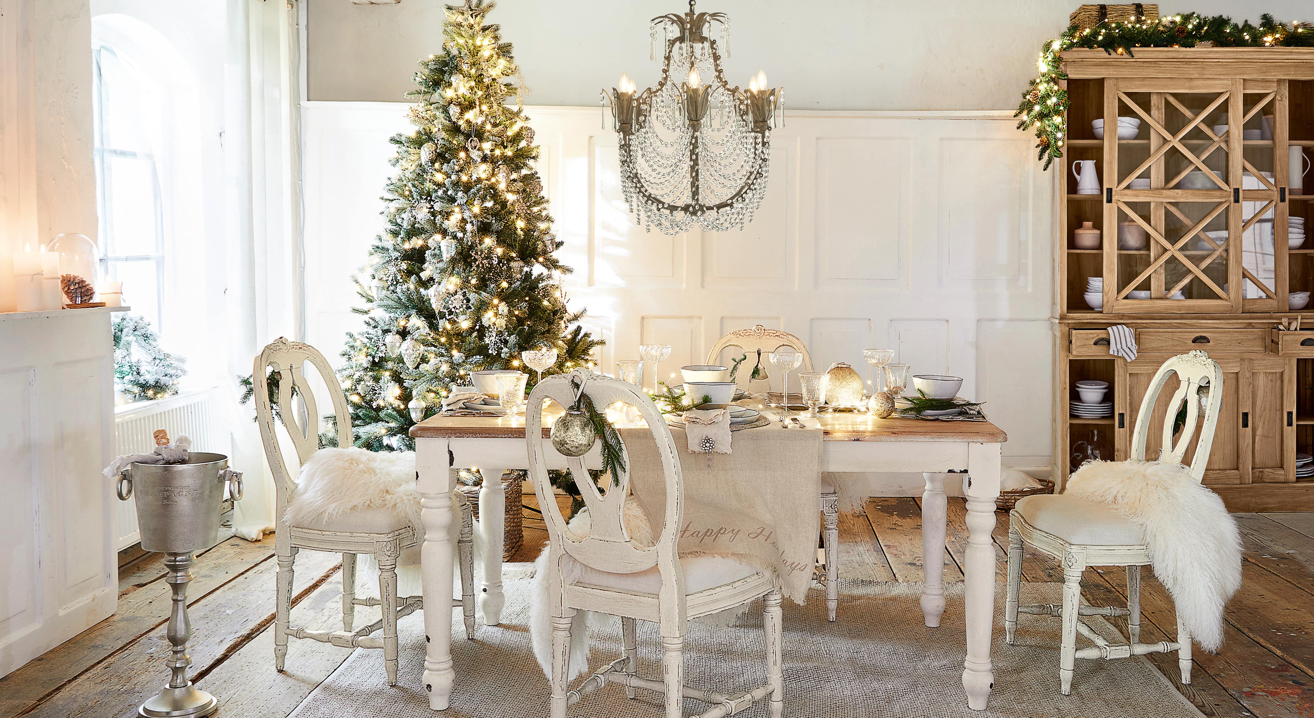 Eetkamer met witte houten tafel en stoelen, feestelijk versierd met kerstboom en kristallen kroonluchter