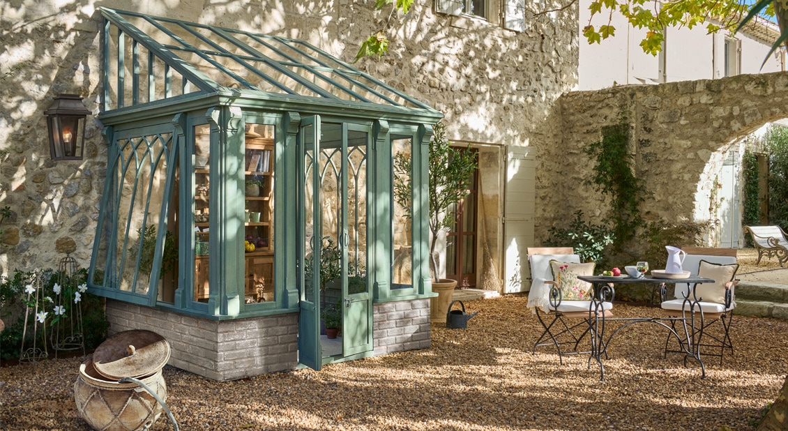 Serre verte en métal sur socle en pierre dans une cour ensoleillée avec mobilier de jardin en fer forgé