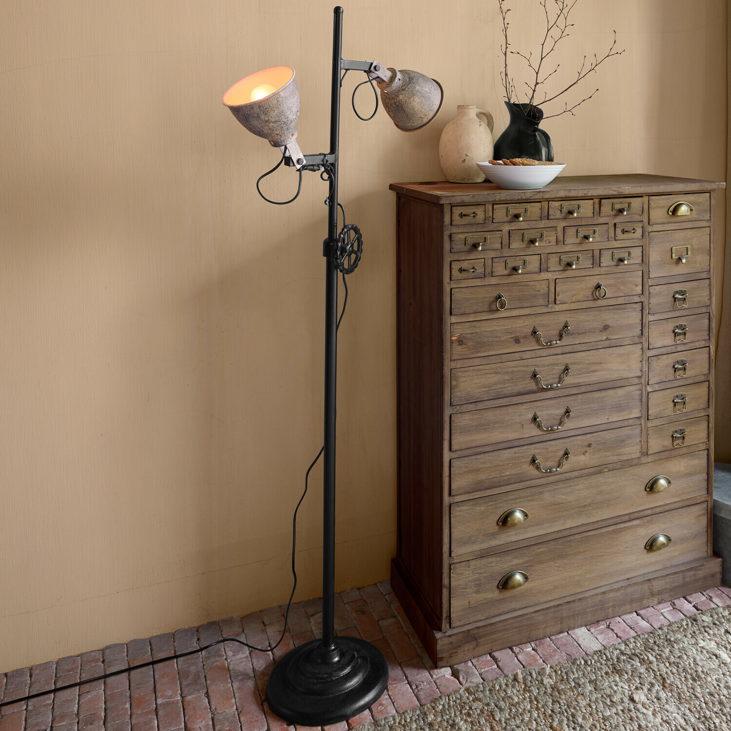Vloerlamp Fenric Staande lamp met grijze metalen kappen en zwarte voet naast een rustieke houten ladekast