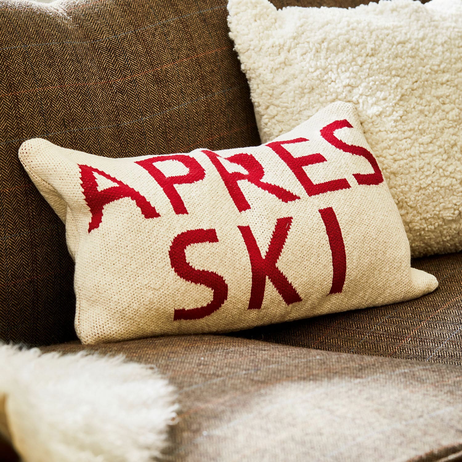 Beige kussen met rode tekst Apres Ski op bruine bank met zachte bontdeken