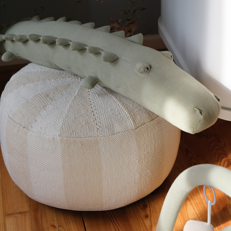 Crocodile en peluche vert clair posé sur un pouf beige tissé sur un sol en bois, décoration d'intérieur enfantine